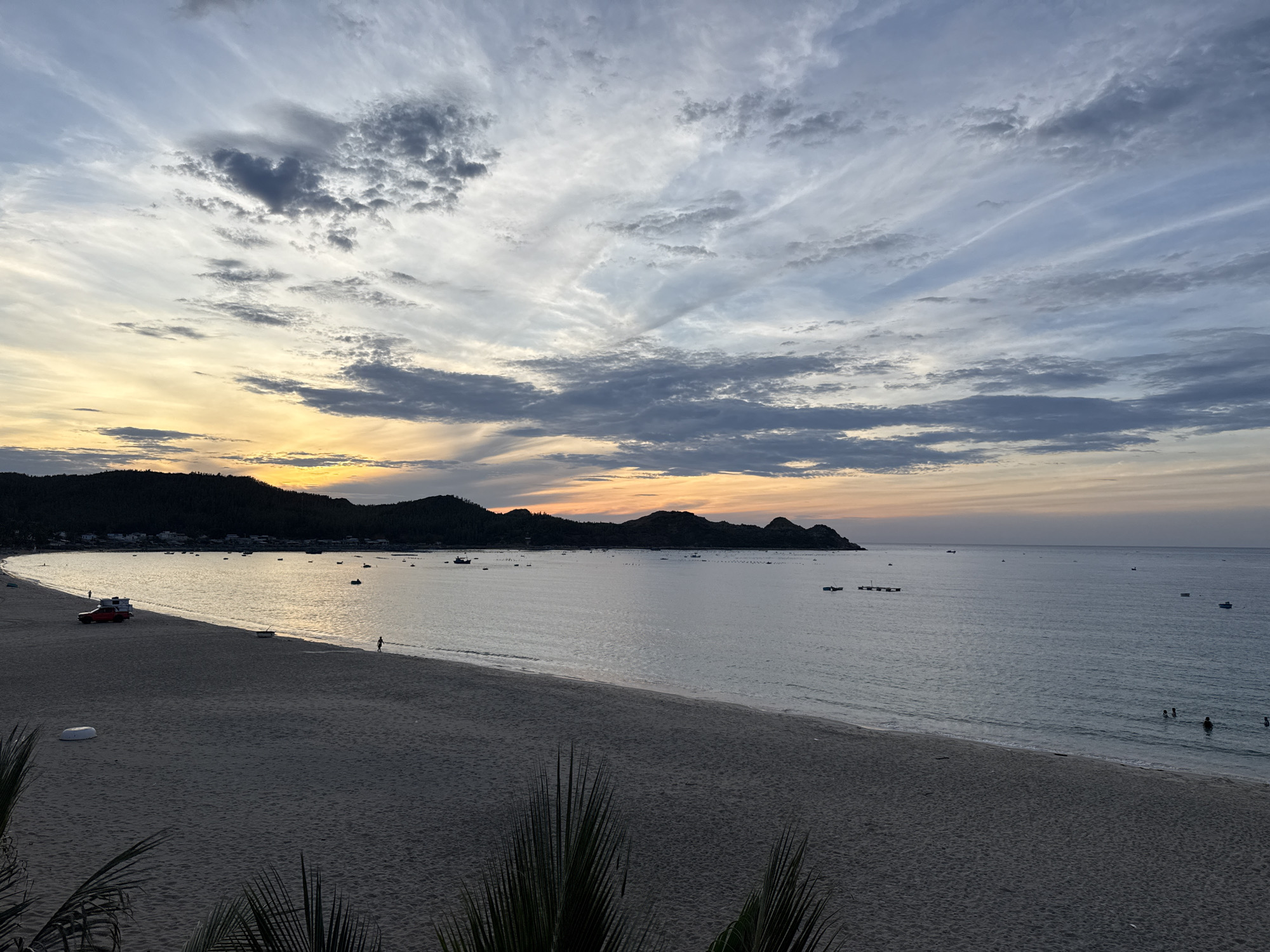 This is a panoramic, eye-level, outdoor photograph of a beach at sunset in Lệ Uyên, Vietnam. The sky is filled with dramatic clouds in shades of grey, white, and light blue, with streaks of yellow and orange near the horizon, indicating the setting sun. The sea is calm, reflecting the colors of the sky. The beach is wide and sandy, with a few palm fronds in the foreground obscuring the very bottom of the frame. In the distance, a dark, hilly coastline is visible, dotted with buildings. Several small boats are scattered across the water. On the beach, a red vehicle with a trailer is parked near the water's edge, and a few people are visible, including one walking on the shore and several others swimming in the sea. The overall scene is serene and tranquil.