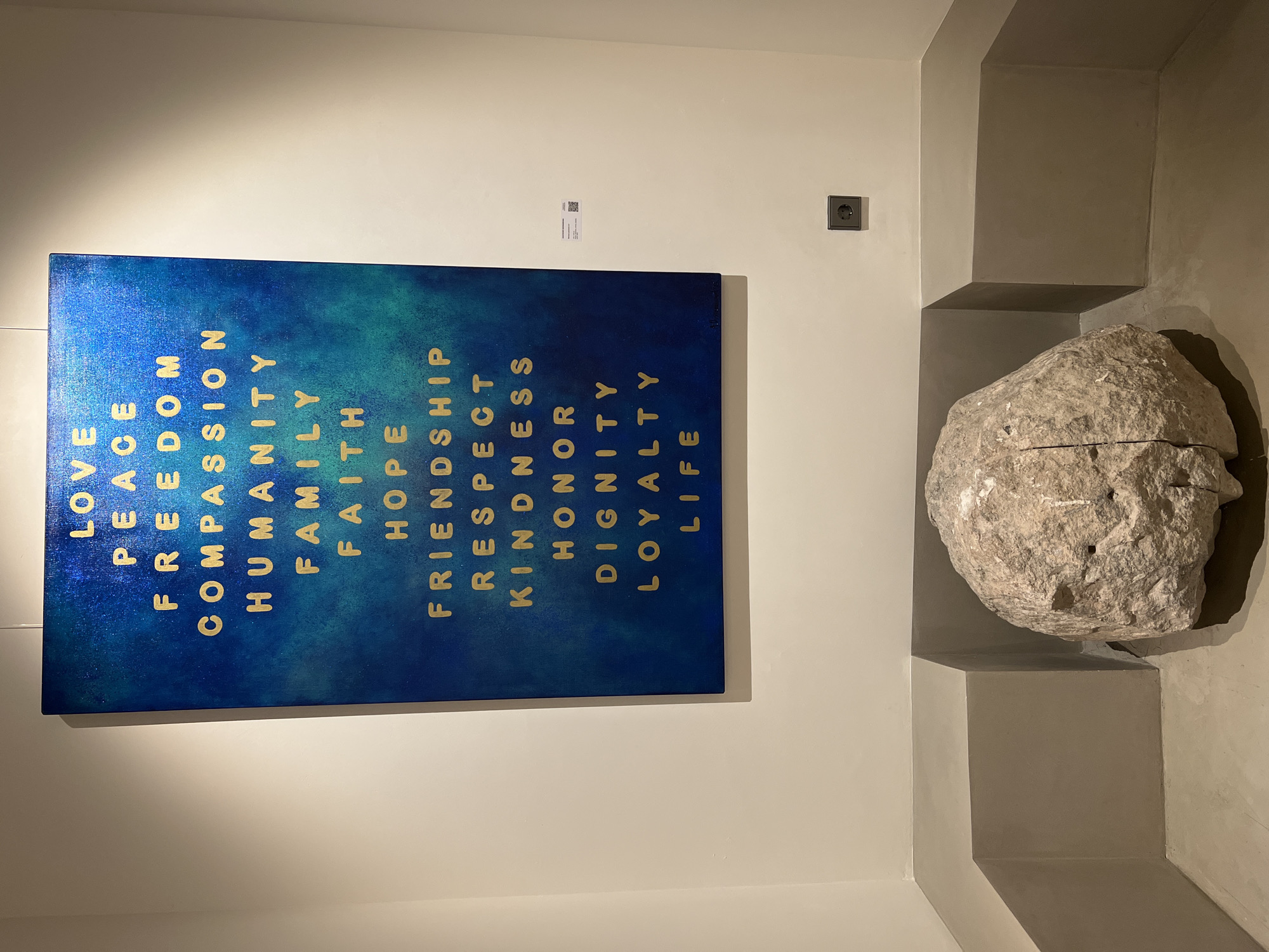 A vertical, eye-level shot depicts a wall in an art gallery or museum, with a blue painting on the left and a large stone sculpture on the right. The painting features a gradient blue background with words like "LOVE," "PEACE," "FREEDOM," "COMPASSION," "HUMANITY," "FAMILY," "FAITH," "HOPE," "FRIENDSHIP," "RESPECT," "KINDNESS," "HONOR," "DIGNITY," "LOYALTY," and "LIFE" in golden, vertically-aligned letters. A small, rectangular label is on the wall above the painting and to the right, and an electrical outlet is visible further to the right. The stone sculpture rests on a stepped architectural structure. The wall is painted a neutral color. No people are visible. The photograph does not contain sufficient information to determine the location as Moscow, Russia.