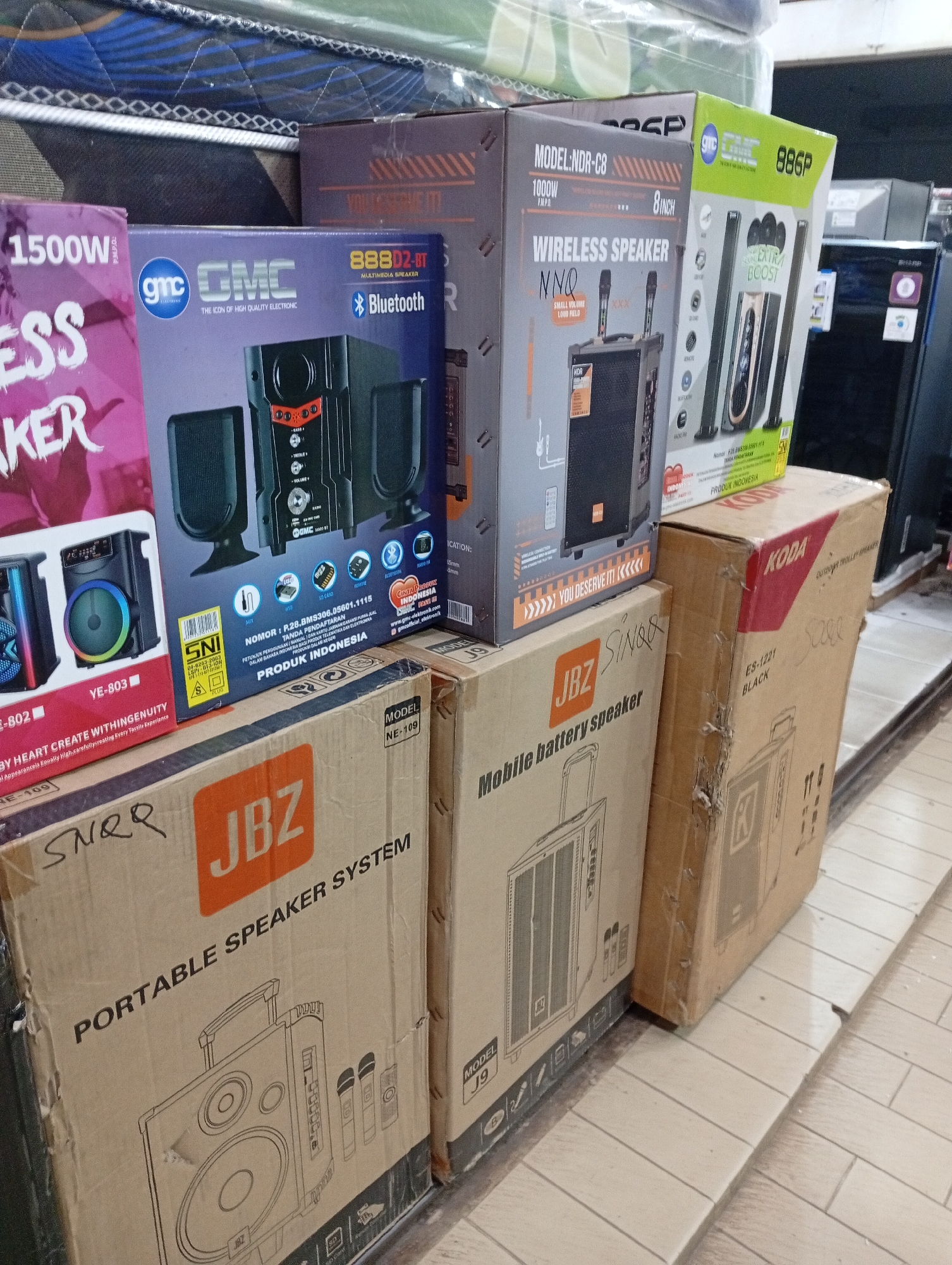 The image shows a retail display of boxed audio equipment, likely in a store in Batam, Indonesia. In the foreground, several boxes are stacked, featuring brands like "JBL", "GMC", and "KODA". The boxes display images and specifications of speakers and audio systems. Behind these, more boxes are visible, including some with "1500W" and "ESSAKER" branding. In the background, a large appliance, possibly a refrigerator, is partially visible. The flooring appears to be tiled. The scene is illuminated by artificial lighting.