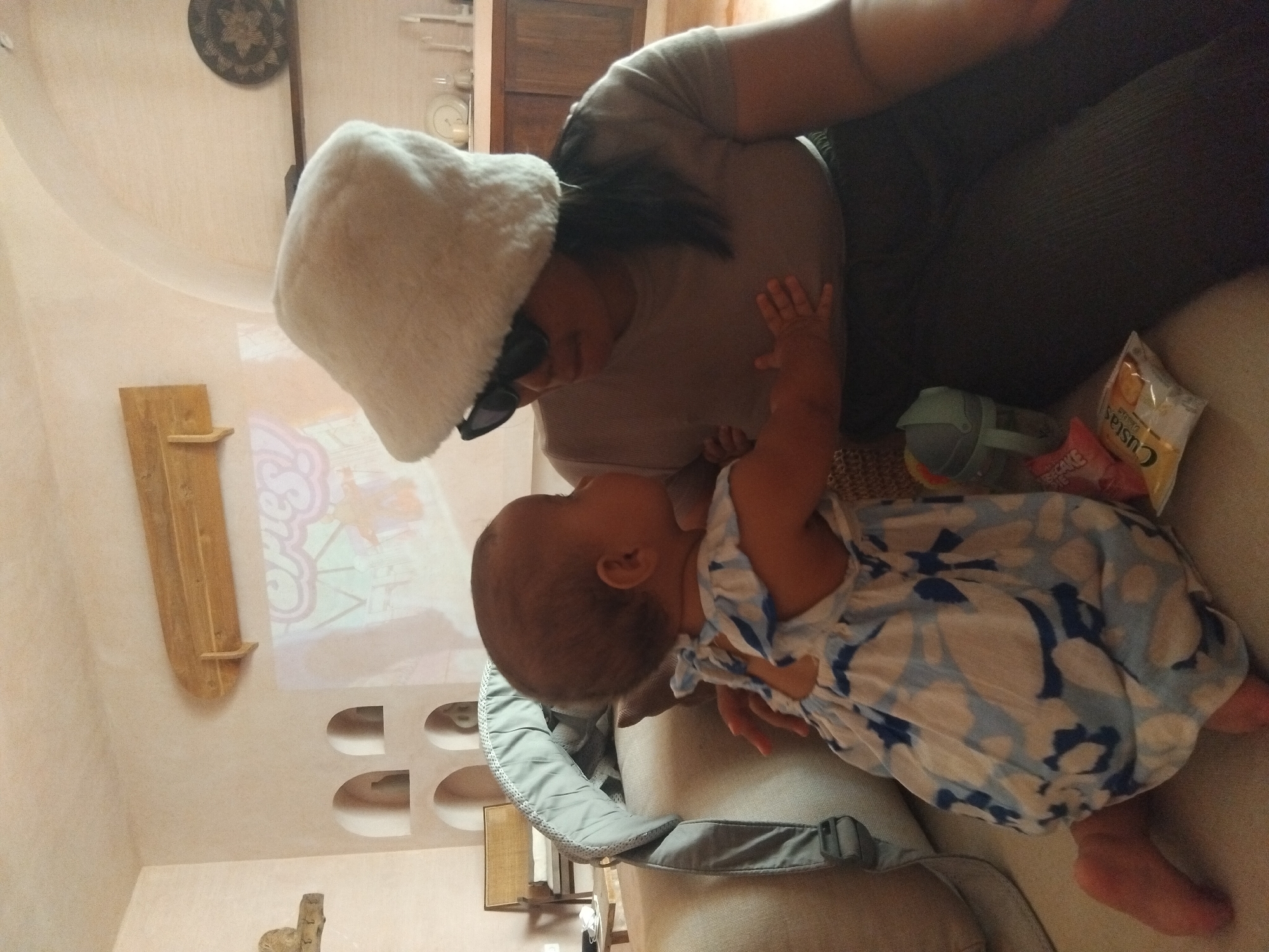 The image shows a woman wearing a white furry hat and sunglasses, leaning towards a baby in a blue and white patterned dress. The baby is lying on a light grey baby carrier placed on a beige couch. Next to the baby, there is a snack bag and a small toy. The background includes a wall with decorative recesses and a shelf, and an image is projected on the wall. The location is Unknown, Unknown.