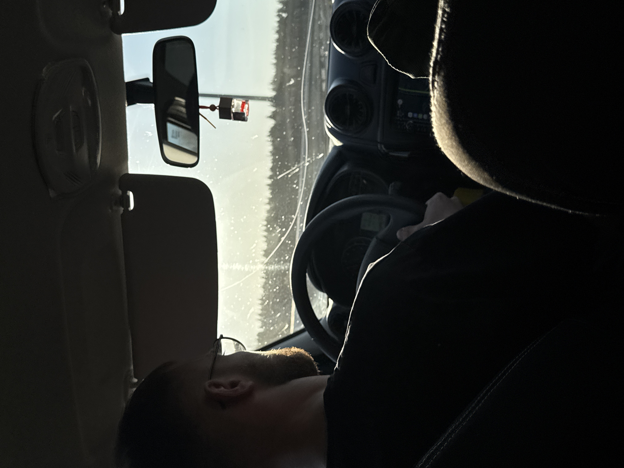 The image depicts the interior of a vehicle, possibly a Jeep, with a driver in view. The driver, a male with glasses and a beard, is partially visible in the lower-left corner. His hand is visible on the steering wheel. The dashboard and front windshield are visible. A rearview mirror hangs above the windshield. Outside the car, a light-colored sky and what appears to be a forested area are visible. No location specific to the city of Yanychi, Russia is discernable.