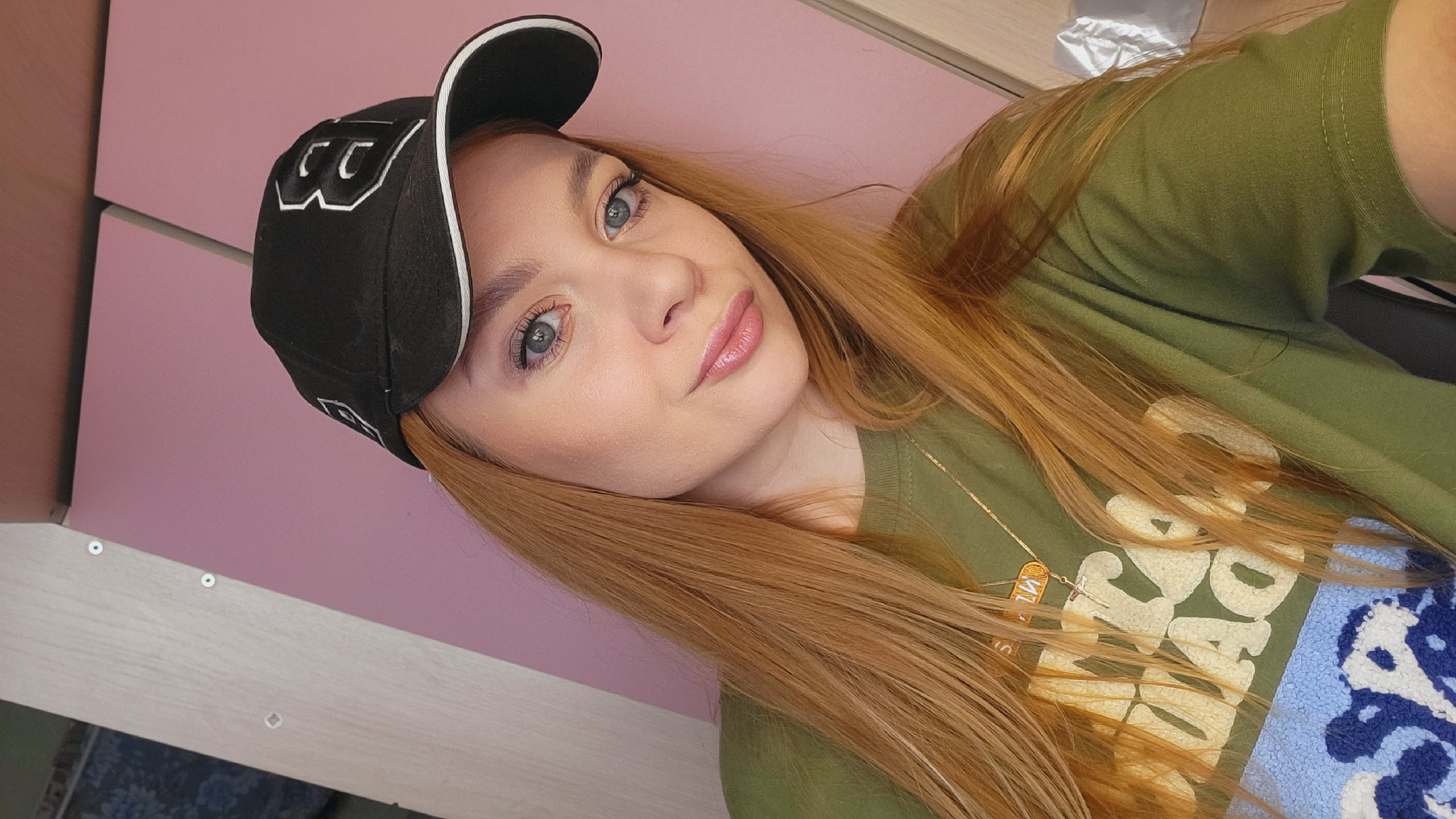 A woman with long, strawberry-blonde hair, light skin, and light eyes is looking at the camera. She is wearing a black baseball cap with white trim and a logo, and an olive green t-shirt with a graphic design. A delicate gold necklace is visible around her neck. The background consists of a pink wall and a white surface. The location information is not apparent in the image.