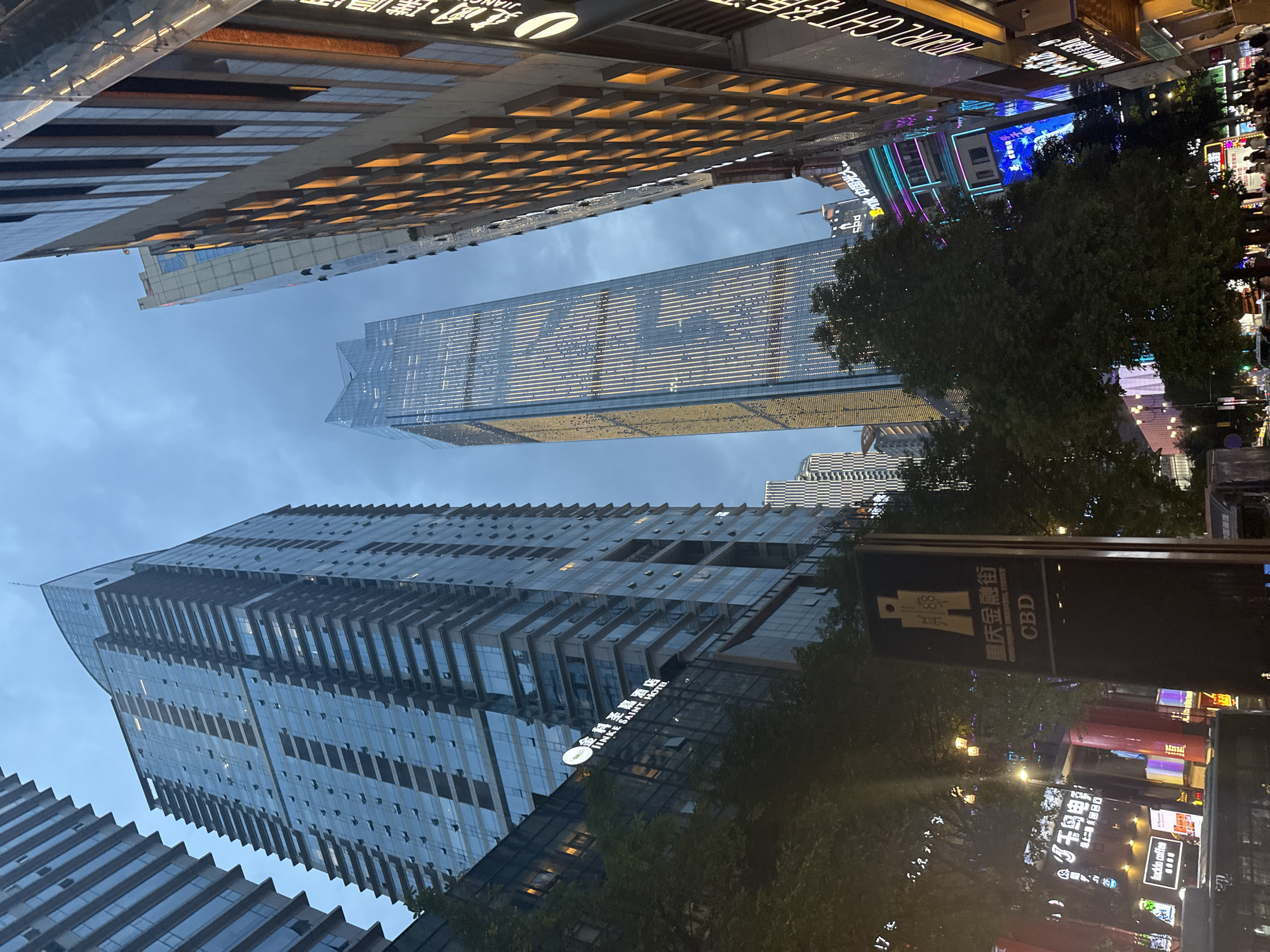 The image captures a low-angle view of a bustling street scene in the Yuzhong District of Chongqing, China, at dusk. Towering skyscrapers, characteristic of a modern city, dominate the skyline, their glass facades reflecting the dimming sky and the illuminated windows of other buildings. The building on the left, featuring a distinctive curved top, is a tall, modern high-rise with many visible windows. To its right, another slender skyscraper with a pointed top is illuminated with vertical lines of light, creating a striking visual effect. In the foreground, a prominent black sign with gold Chinese characters and the English acronym "CBD" stands near a streetlamp and lush green trees, indicating a central business district. Further down the street, businesses with illuminated signage, including "Luckin Coffee," are visible, with a few pedestrians walking along the sidewalk. The overall atmosphere suggests a vibrant urban environment transitioning from day to night.