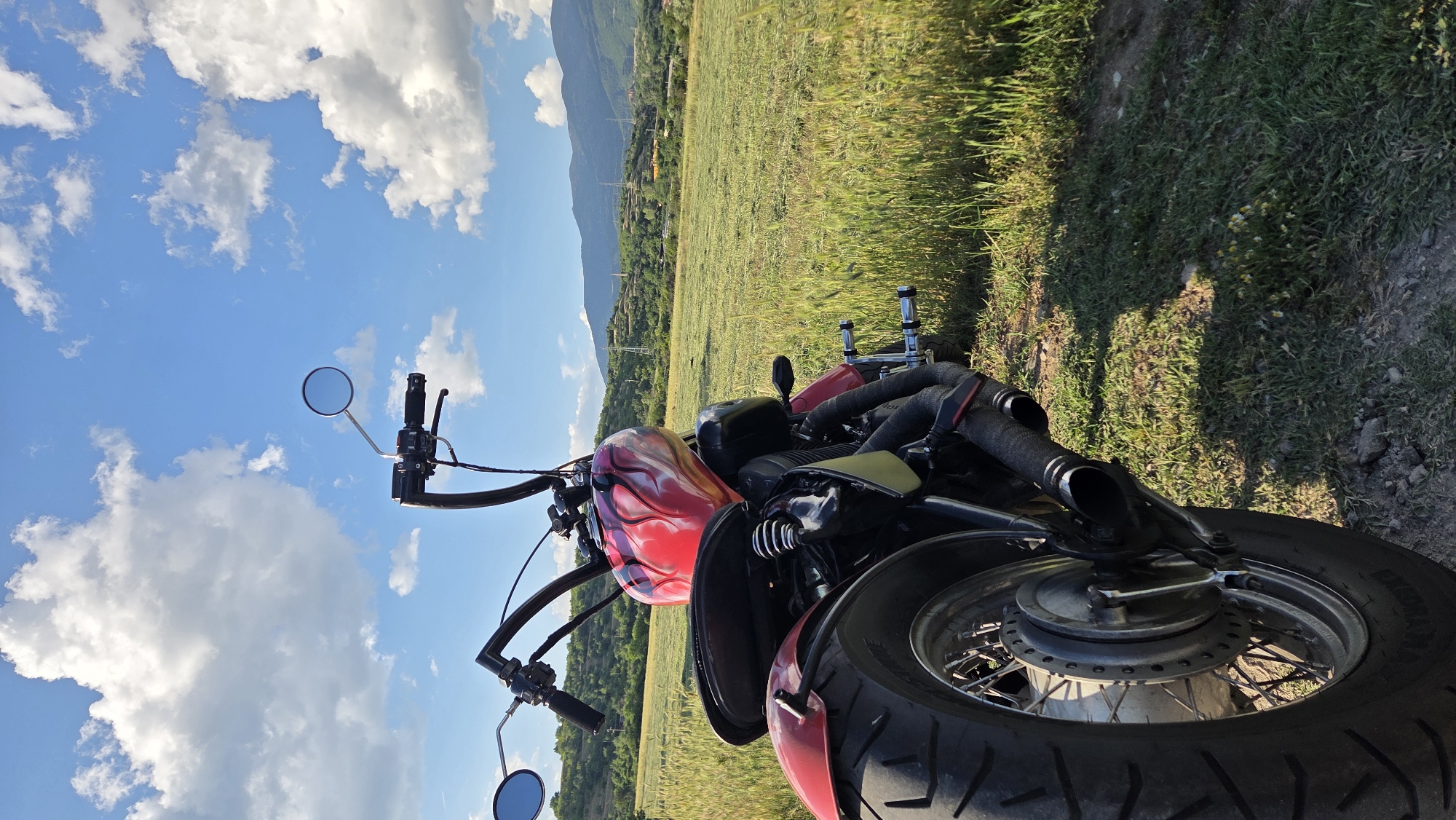 A red and yellow flame-painted motorcycle is parked on a dirt path bordering a grassy field. The motorcycle is viewed from the rear, slightly to the left, with its prominent exhaust pipes and rear tire in focus. Beyond the field, rolling hills dotted with trees and some power lines form the background, under a bright blue sky with scattered white clouds. The scene is set in the Rila region of Bulgaria, evidenced by the natural landscape. There are no people visible in the image.
