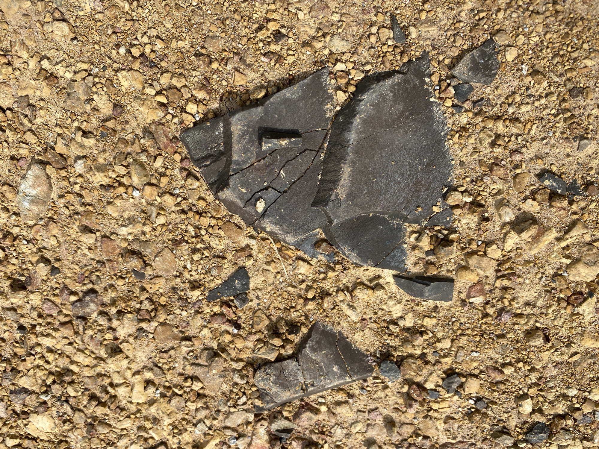 A close-up, top-down view shows dark, fragmented rock pieces scattered on a sandy, gravelly ground. The rock fragments are angular and appear to have sharp edges, with one large piece in the center exhibiting visible cracks and a slightly raised protrusion. The surrounding ground is composed of a mixture of small, irregularly shaped pebbles and sand, varying in color from light tan to reddish-brown and grey. The lighting suggests an outdoor environment, casting subtle shadows on the ground. There are no people or discernible structures visible in the image. The location is the city of Ko Siboya, Thailand.