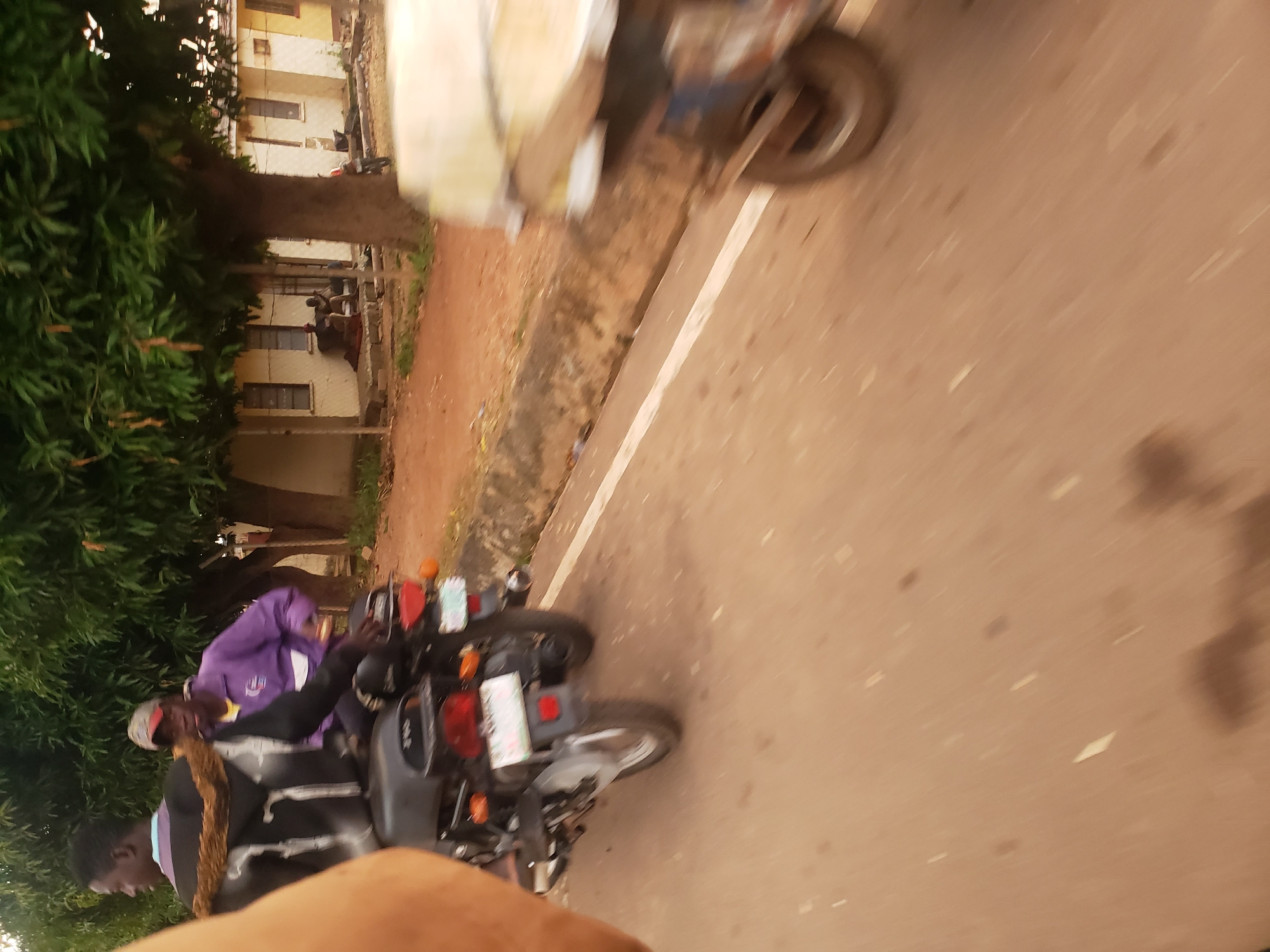 The image shows a scene in Narayi, Nigeria, capturing two individuals on a motorcycle. The person in the foreground, seated behind the driver, wears a dark jacket with a fur-lined collar and a purple garment underneath. The driver is wearing a purple jacket and a red cap. The motorcycle, a dark color, has visible license plates and taillights. In the background, a building with several windows and a fence suggests a residential or commercial area. Several trees are present, with one large tree prominent in the middle ground. The road surface is paved, with a white line marking the lane. The image has a slight blur, indicating movement.
