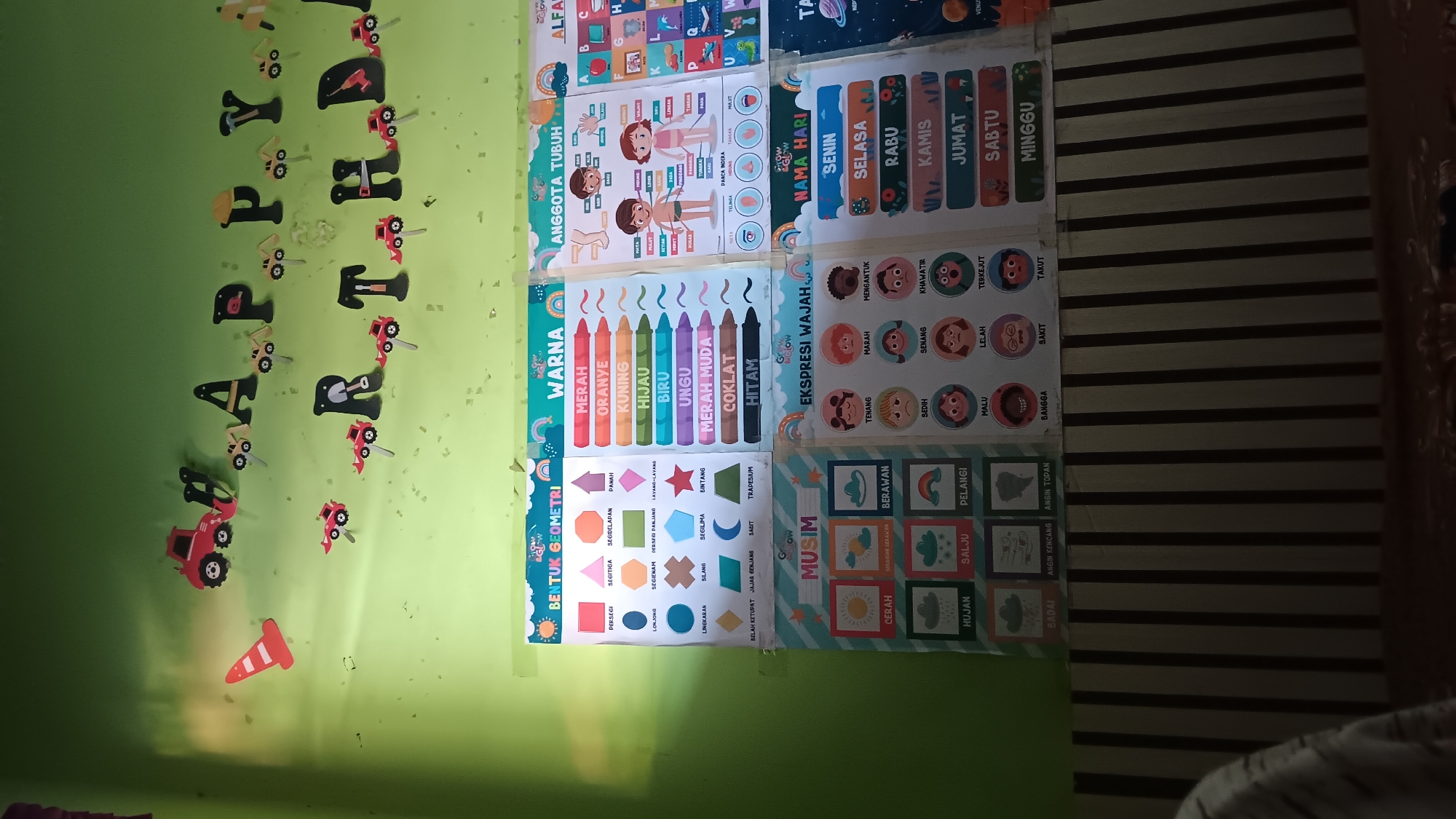The image depicts a wall decorated with colorful educational charts and a "Happy Birthday" banner. The banner, featuring construction vehicle cutouts, is centrally placed and reads "HAPPY BIRTHDAY." Below the banner, on the right side of the image, a chart titled "NAMA HARI" (Name of the Day) is visible, listing the days of the week in Indonesian. To the left of that, a chart titled "EKSPRESI WAJAH" (Facial Expressions) displays various emotions. Further to the left are charts for "MUSIM" (Seasons), "ANGGOTA TUBUH" (Body Parts), "WARNA" (Colors), and "BENTUK GEOMETRI" (Geometric Shapes). The overall environment appears to be an indoor setting, possibly a classroom or a child's room, given the educational content and decorations. The lighting suggests it is daytime. No specific landmarks or elements in the image can confirm the location as Sonobekel, Indonesia.