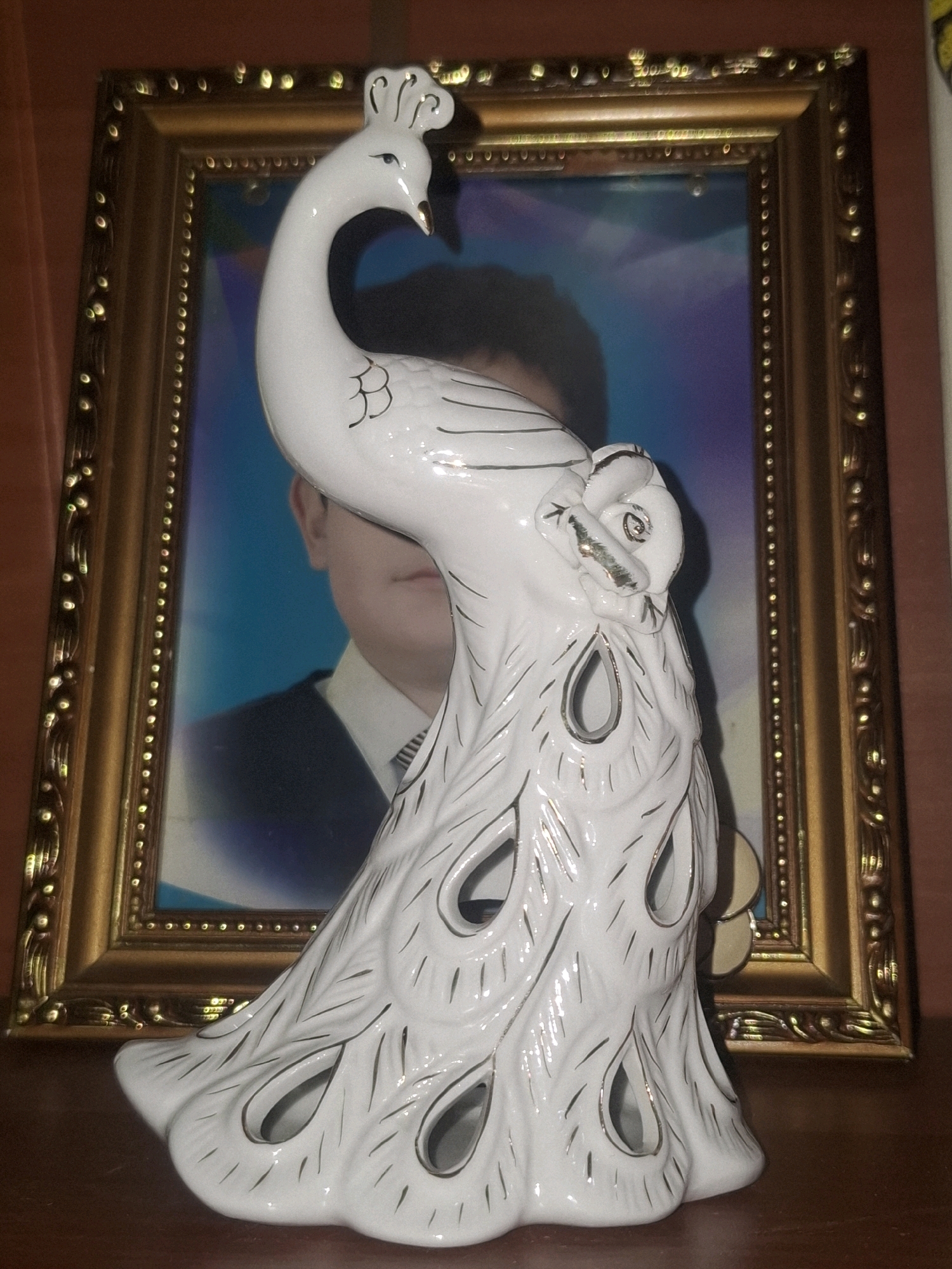 A white ceramic peacock figurine stands in front of a framed photograph. The photograph appears to be a portrait of a person with dark hair and is bordered by a decorative gold frame. The peacock figurine is detailed with black lines and has a long, curved neck with its head turned to the left. Its body is decorated with stylized feather patterns and cut-out shapes that resemble peacock eyes. The figurine is placed on a dark wooden surface, possibly a table or shelf. The background is a reddish-brown wall. This scene is located indoors, in the city of Almaty, Kazakhstan.