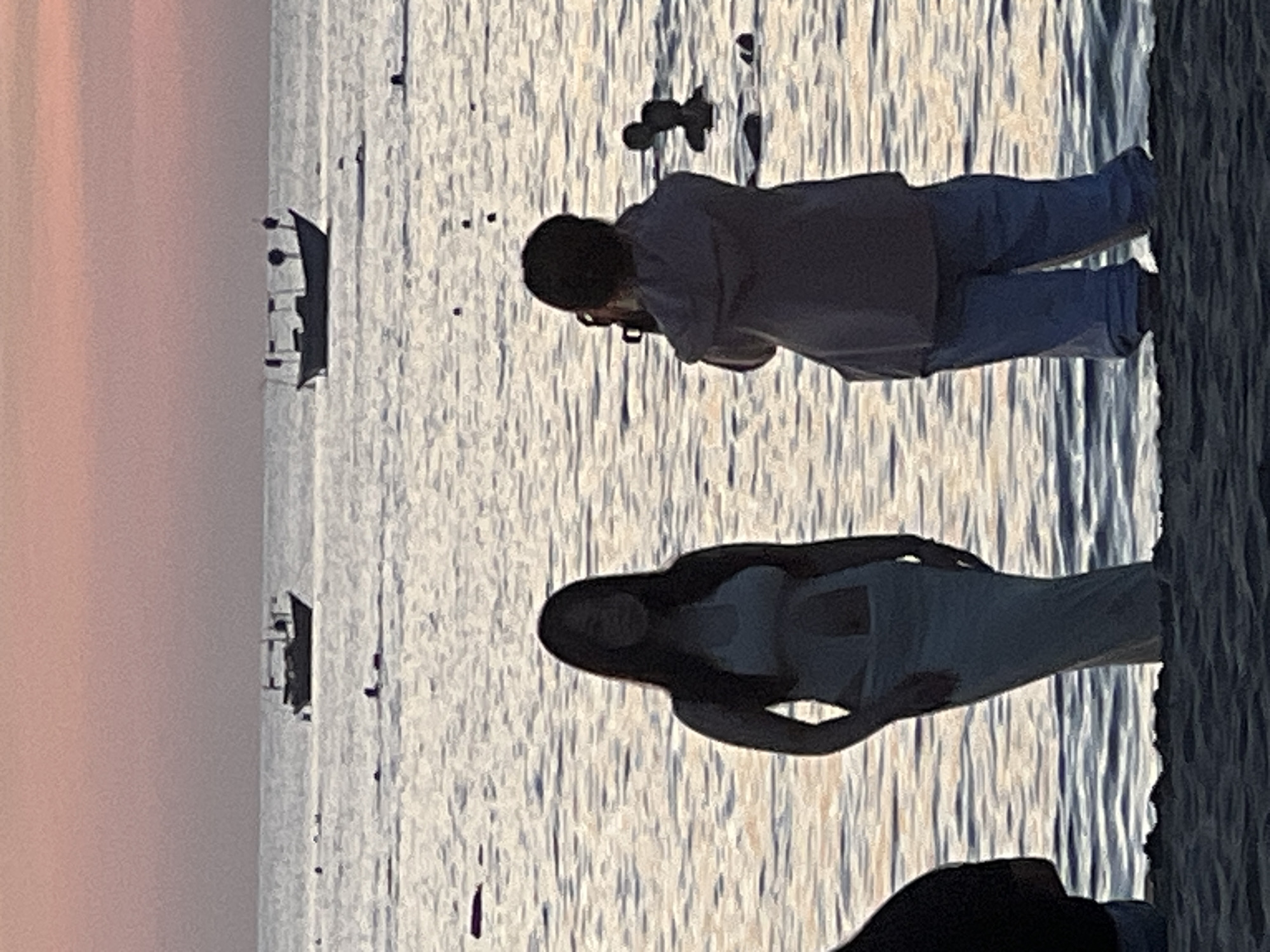 The image depicts a beach scene at dusk, likely in Da Nang, Vietnam, as suggested by the presence of traditional fishing boats. In the foreground, two individuals are partially visible. To the left, a person in dark clothing stands with their back to the viewer. To the right, a person is seen from behind, holding a camera and appearing to take a photograph. They are wearing a light-colored hooded top and blue pants.
In the middle ground, a woman is standing in the shallow water, facing slightly towards the left. She is wearing a light-colored dress with cutouts at the waist. Her silhouette is clearly defined against the water. Further out in the sea, several small fishing boats with distinctive structures are visible. Some people are also seen in the water in the distance, appearing to be swimming or wading. The water reflects the soft hues of the sunset sky, which transitions from a pale pink at the horizon to a muted orange and yellow. The overall atmosphere is calm and serene, characteristic of an evening on a Vietnamese beach.