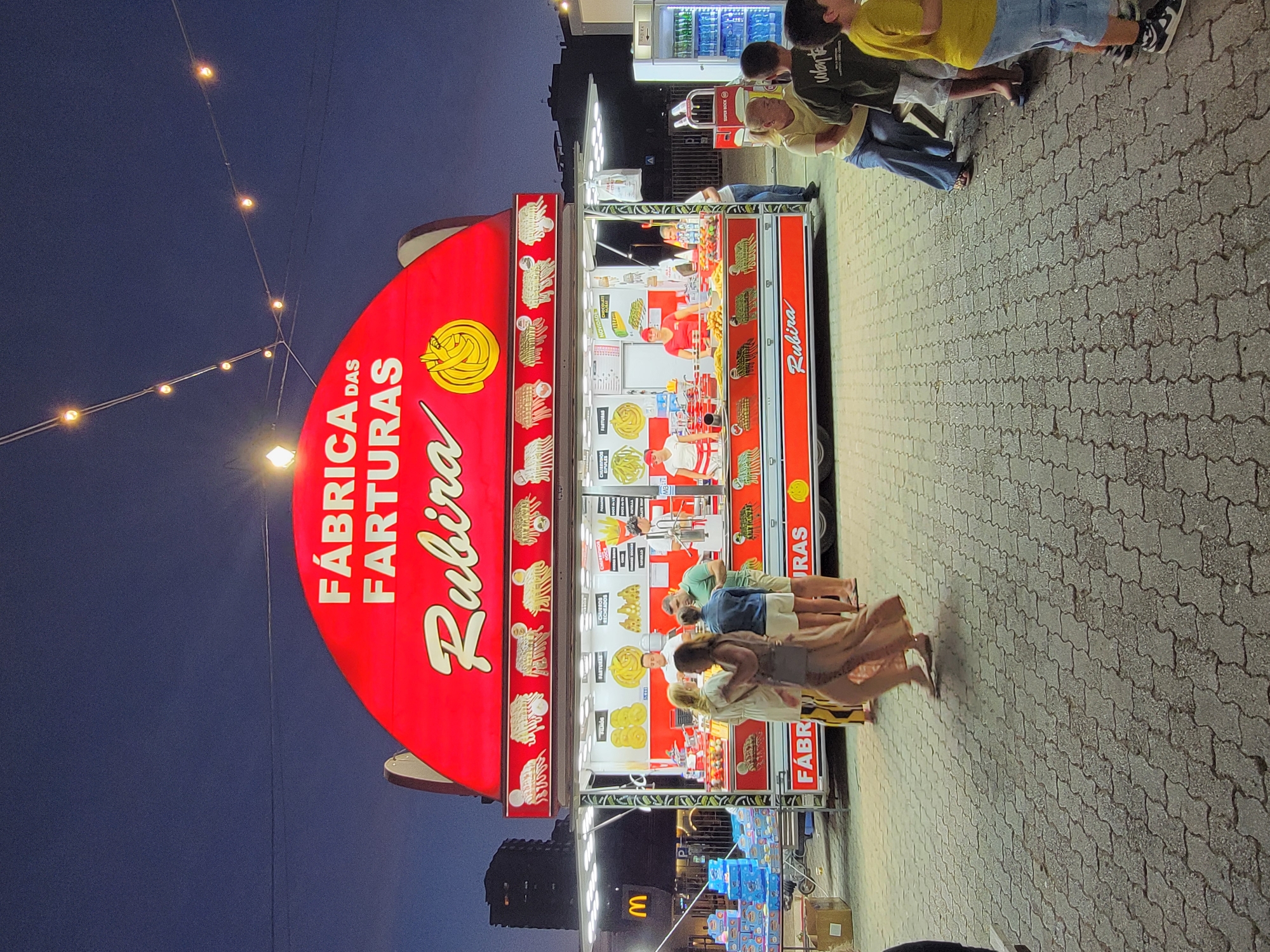 A red food truck, identified as "FÁBRICA DAS FARTURAS Rubira," is parked on a paved area at dusk, likely in Quarteira, Portugal, given the signage and general appearance. The food truck displays images of churros and other fried pastries. Inside the truck, several employees in uniform are preparing food. A few customers are visible, some interacting with the staff and others standing nearby. In the foreground, a group of people, including children, are gathered. String lights are visible overhead, and a building with visible windows is in the background. The ground is paved with interlocking stones. A McDonald's sign is visible in the distance to the left.
