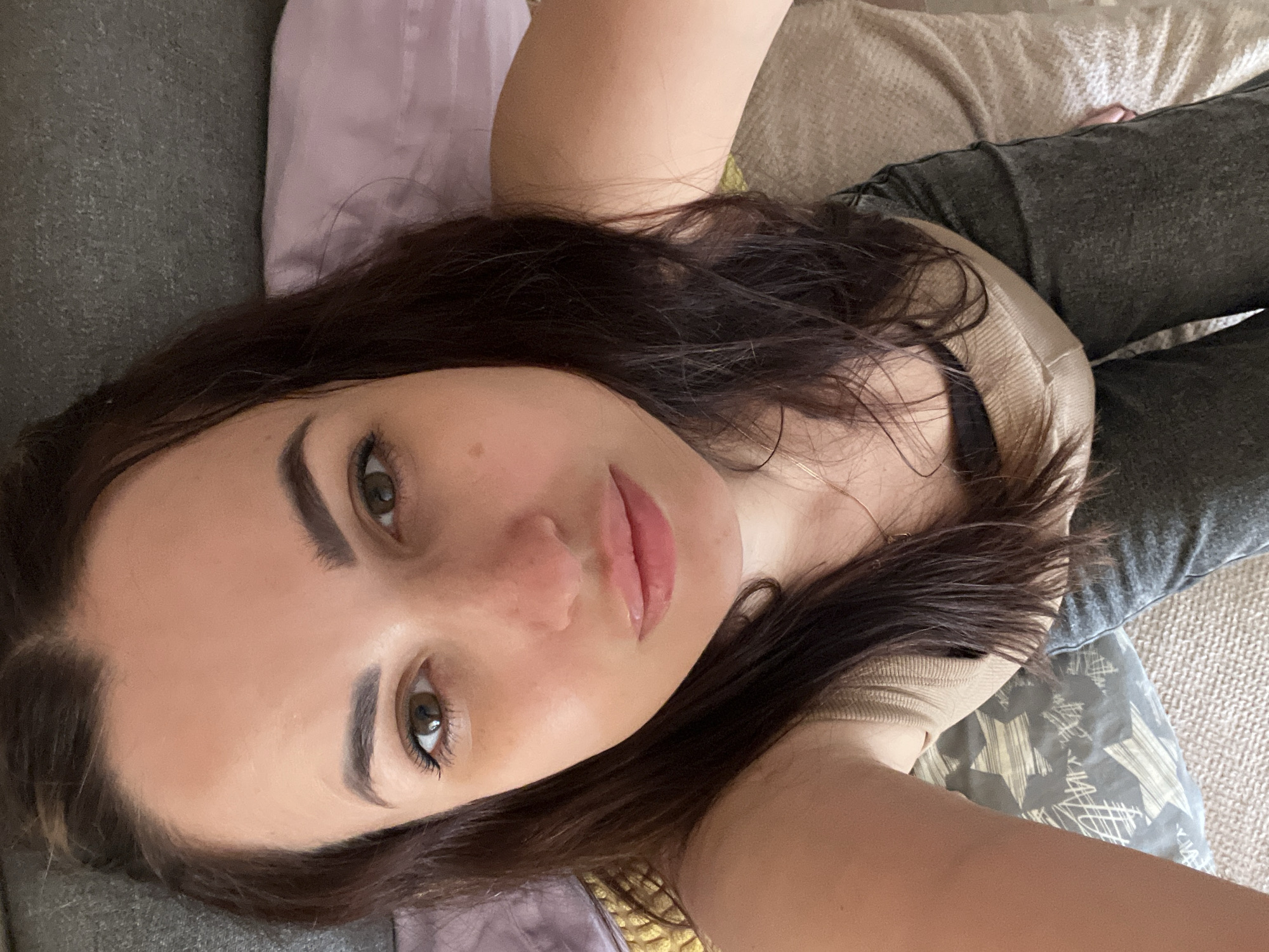 A young woman with dark brown hair and brown eyes is lying down and looking at the camera. She is wearing a beige tank top and dark jeans. Her head is tilted slightly to the left, and her hair is resting on her shoulders and across her face. The background appears to be a bed with pillows and blankets. The lighting is natural and comes from the front. The image is a selfie taken from a high angle.