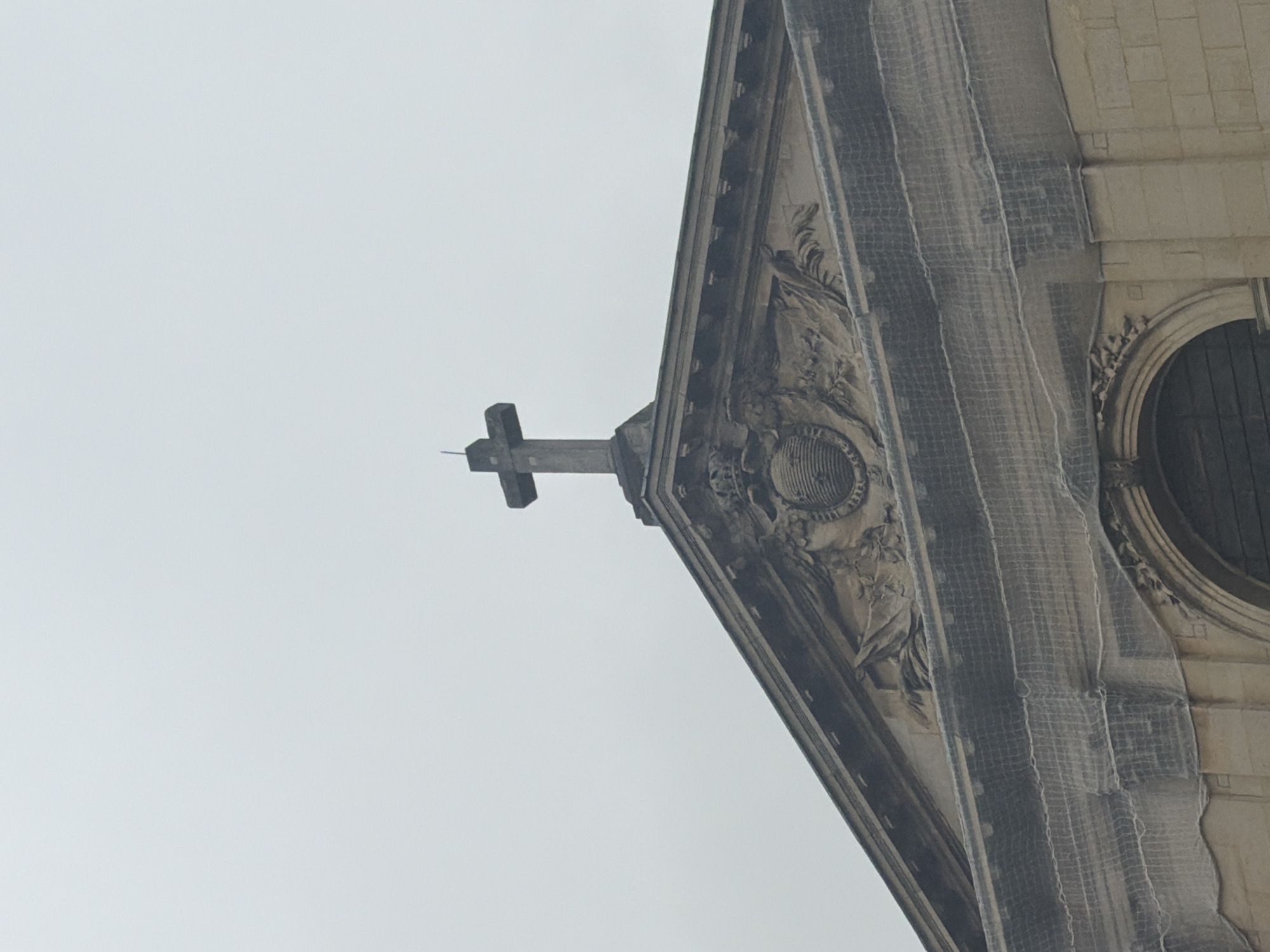 An eye-level shot shows a stone building with architectural details in Paris, France. The building, angled slightly, features an ornate pediment adorned with foliage and a central shield-like emblem. A simple stone cross extends outwards from the pediment's apex. The building is under maintenance, indicated by a mesh-like construction tarp partially covering the side of the building, partially obscuring the corner edge and a circular window on the right side. The sky is overcast, creating a soft, diffused light across the scene.