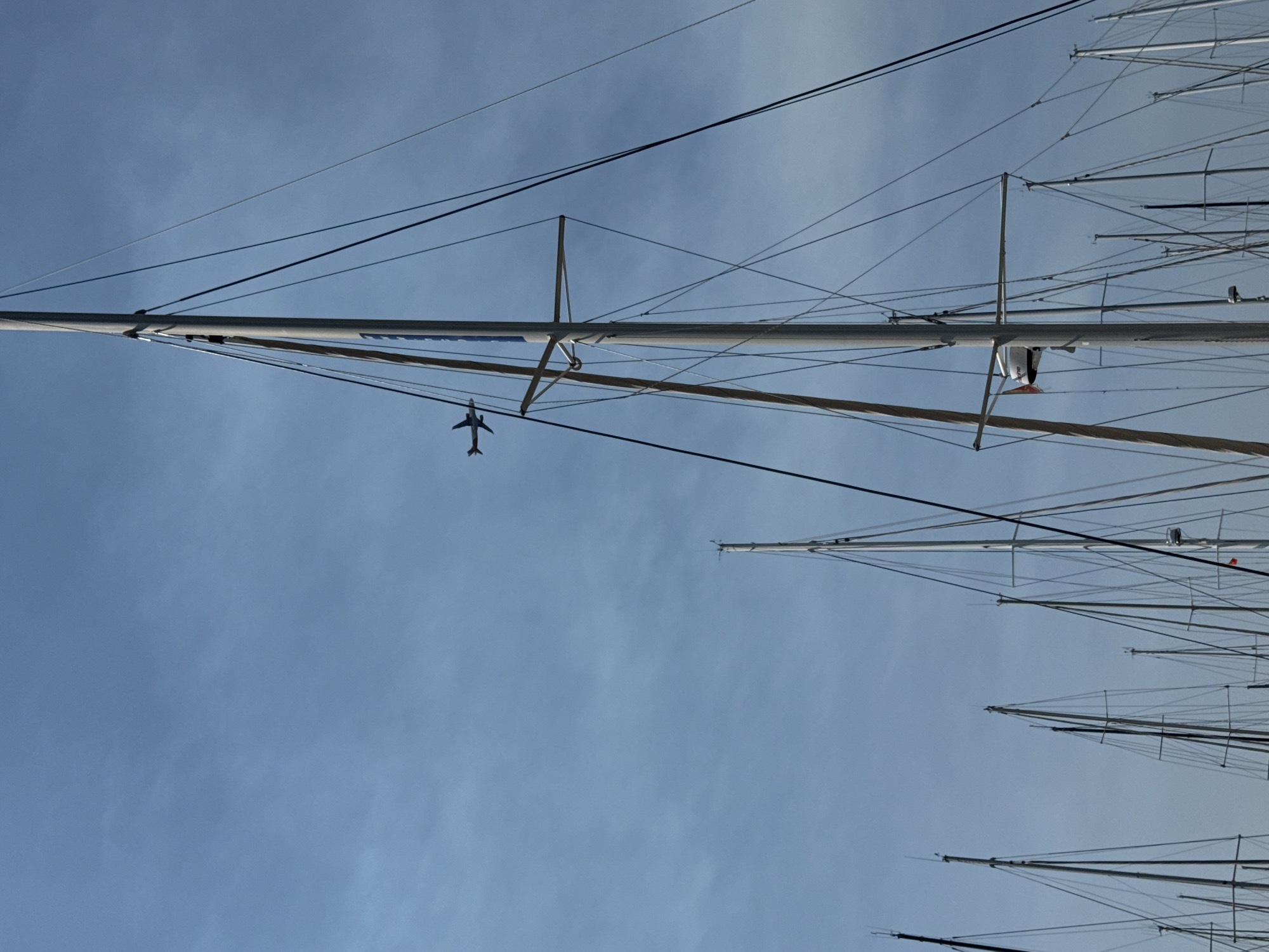 An airplane is visible in the sky, flying above a harbor in Tivat, Montenegro. The airplane is silhouetted against the blue sky. The foreground of the image is dominated by the masts and rigging of several sailboats. The sailboats are docked and their masts create a complex geometric pattern against the sky. The sky is a clear blue with some wispy clouds. The overall scene depicts a peaceful harbor on a sunny day.