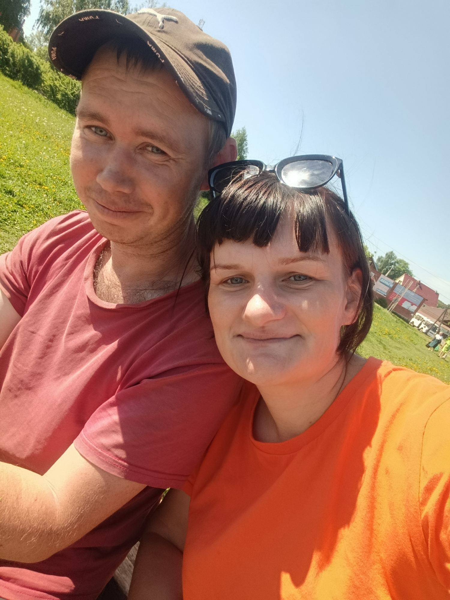 Two people, a man and a woman, are posing for a selfie outdoors in Фоки, Russia. The man is wearing a dark baseball cap and a reddish-pink t-shirt. The woman is wearing an orange t-shirt and has sunglasses on her head. A grassy hill is in the background. In the distant background, there are buildings, including a red-roofed structure, and vehicles, possibly buses. There are also some people in the distance.