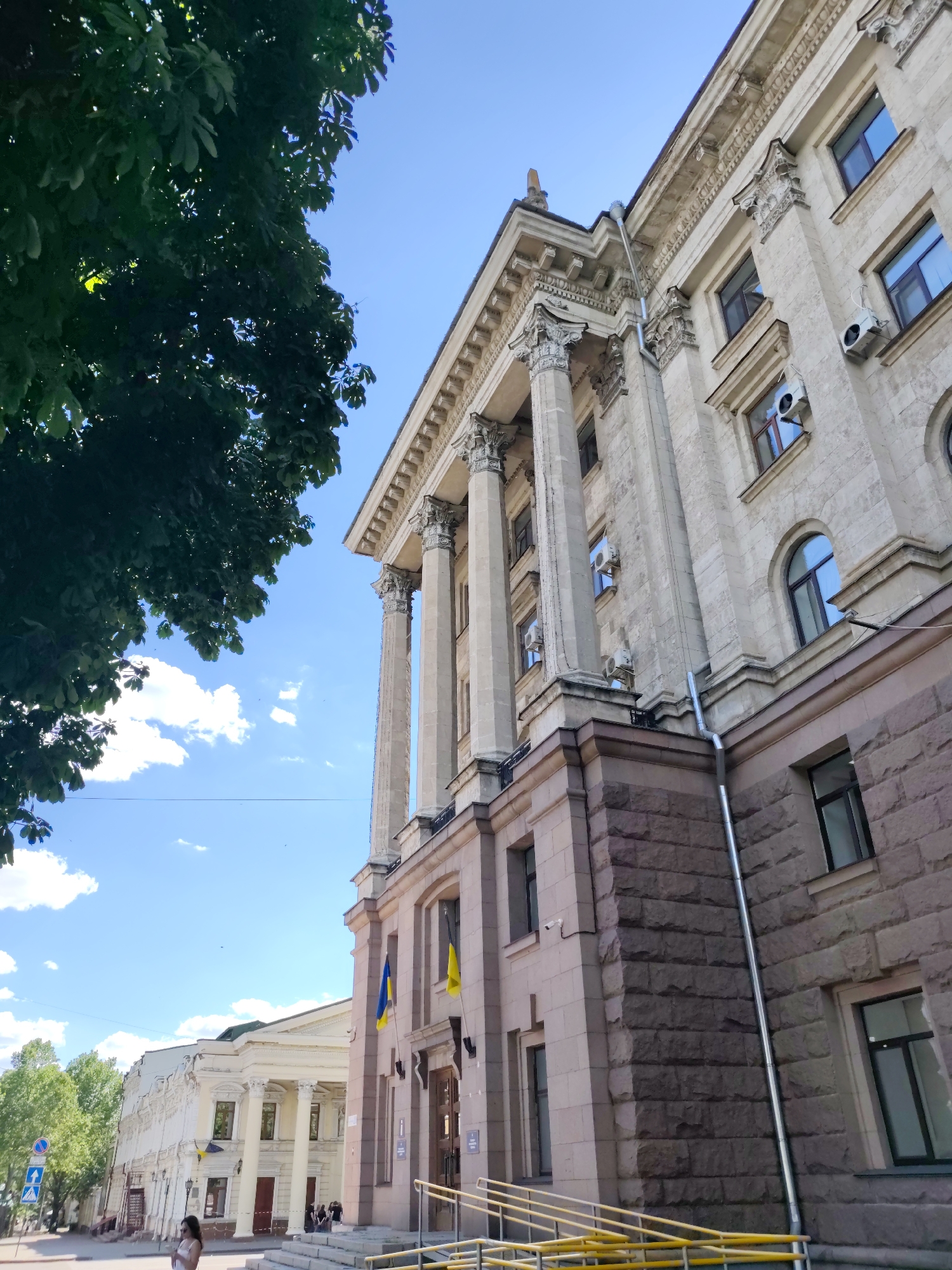 The image depicts the facade of a large, ornate building in Mykolaiv, Ukraine. The architecture features classical elements such as prominent columns and decorative cornices. The building is constructed with light-colored stone on the upper floors and a darker, rough-hewn stone base. Several windows with dark frames are visible, and air conditioning units are mounted outside some of them. Ukrainian flags, blue and yellow, are displayed on poles attached to the building.
In the foreground, a tree with dense green foliage partially obscures the view of the sky and the building. The sky is a clear blue with scattered white clouds. In the background, another building with similar architectural styling is visible, also adorned with Ukrainian flags. A woman in a light-colored dress stands on a sidewalk near the background building, looking towards the right of the frame. She is positioned near a traffic sign and a yellow metal railing. Yellow railings are also present in the foreground, leading up to the steps of the main building. The overall scene suggests a bright, daytime setting in an urban environment.