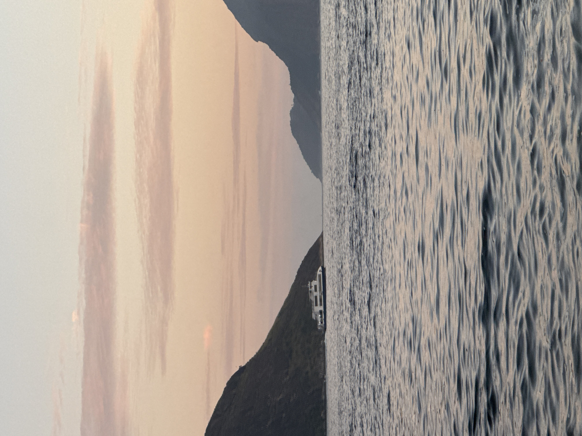 This image captures a serene seascape with a ferry sailing on calm waters under a soft, pastel-colored sky at dusk. The scene appears to be located in Guanabara Bay, with the iconic mountains of Niterói, Brazil, forming a dramatic backdrop. The ferry, a white vessel with blue trim, is positioned in the mid-ground, leaving a gentle wake behind it. The water is a deep blue-grey, reflecting the muted tones of the sky. Wispy clouds with hints of pink and orange streak across the horizon, adding to the tranquil atmosphere. The silhouette of a second, more distant mountain range can be seen to the right, partially obscuring the setting sun. The overall impression is one of peacefulness and natural beauty, characteristic of the coastal landscapes of Rio de Janeiro's neighboring city.