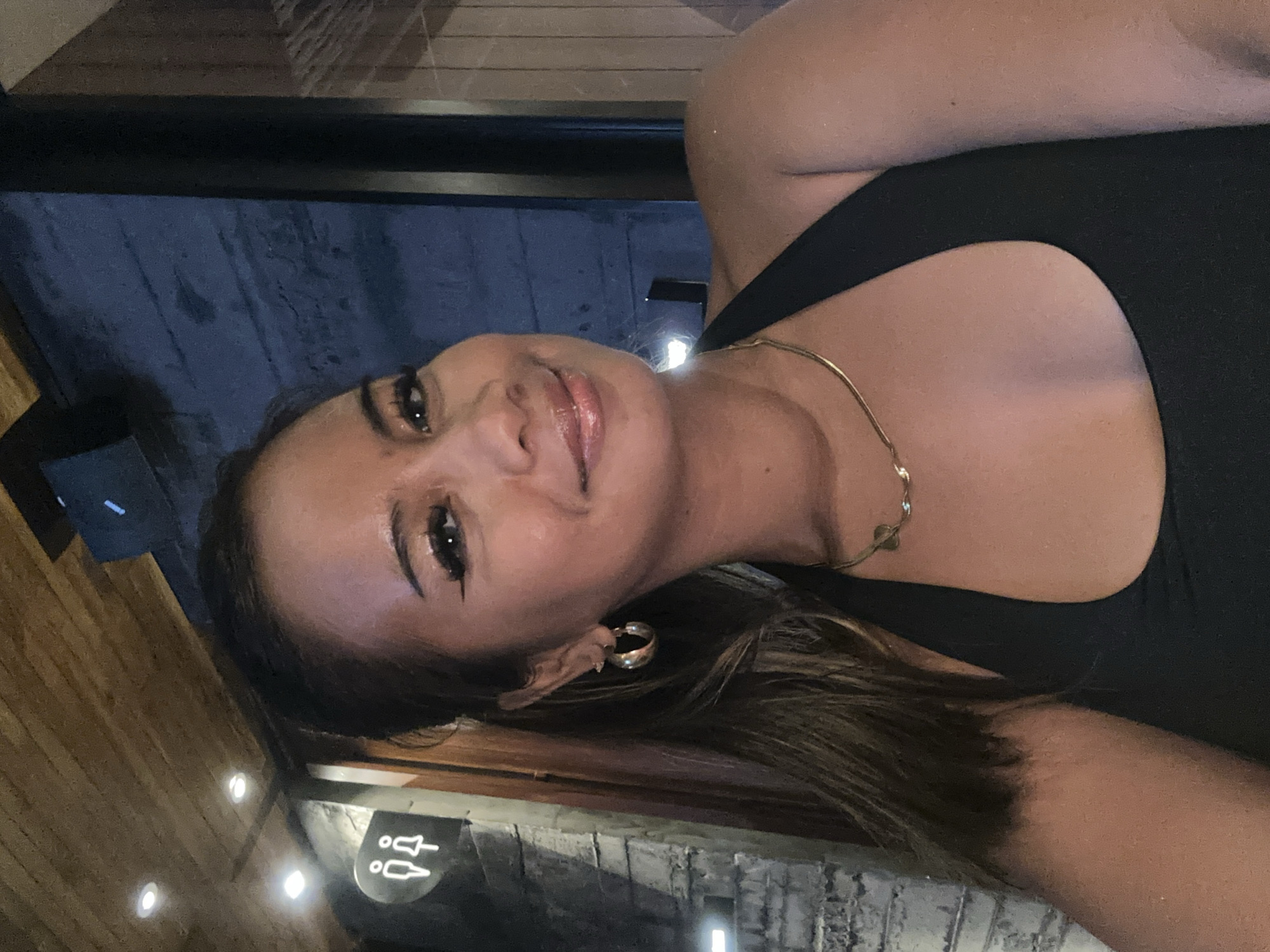 A young woman is captured in a close-up selfie, her head tilted to the right, showcasing a warm smile and glossy lips. She has long, dark brown hair, dark eyes with noticeable eyeliner and mascara, and a natural complexion. She is wearing a black sleeveless top, a delicate gold chain necklace with a small, round pendant, and a thick gold hoop earring on her visible left ear.
The setting appears to be indoors at an establishment, likely a restaurant, bar, or club, given the dim lighting and background elements. To the bottom-left of the frame, there's a wooden paneled wall with several bright, circular recessed lights. To the bottom-right, a textured wall, possibly dark brick or stone, is visible. Behind her head and shoulders, a dark, possibly blue, surface is present.
A distinct white sign is visible in the lower-left, featuring stick figures of a male and a female, indicating the location of restrooms. The woman's expression suggests happiness and confidence, as she looks directly at the camera. The overall ambiance is relaxed and social, characteristic of an evening outing.