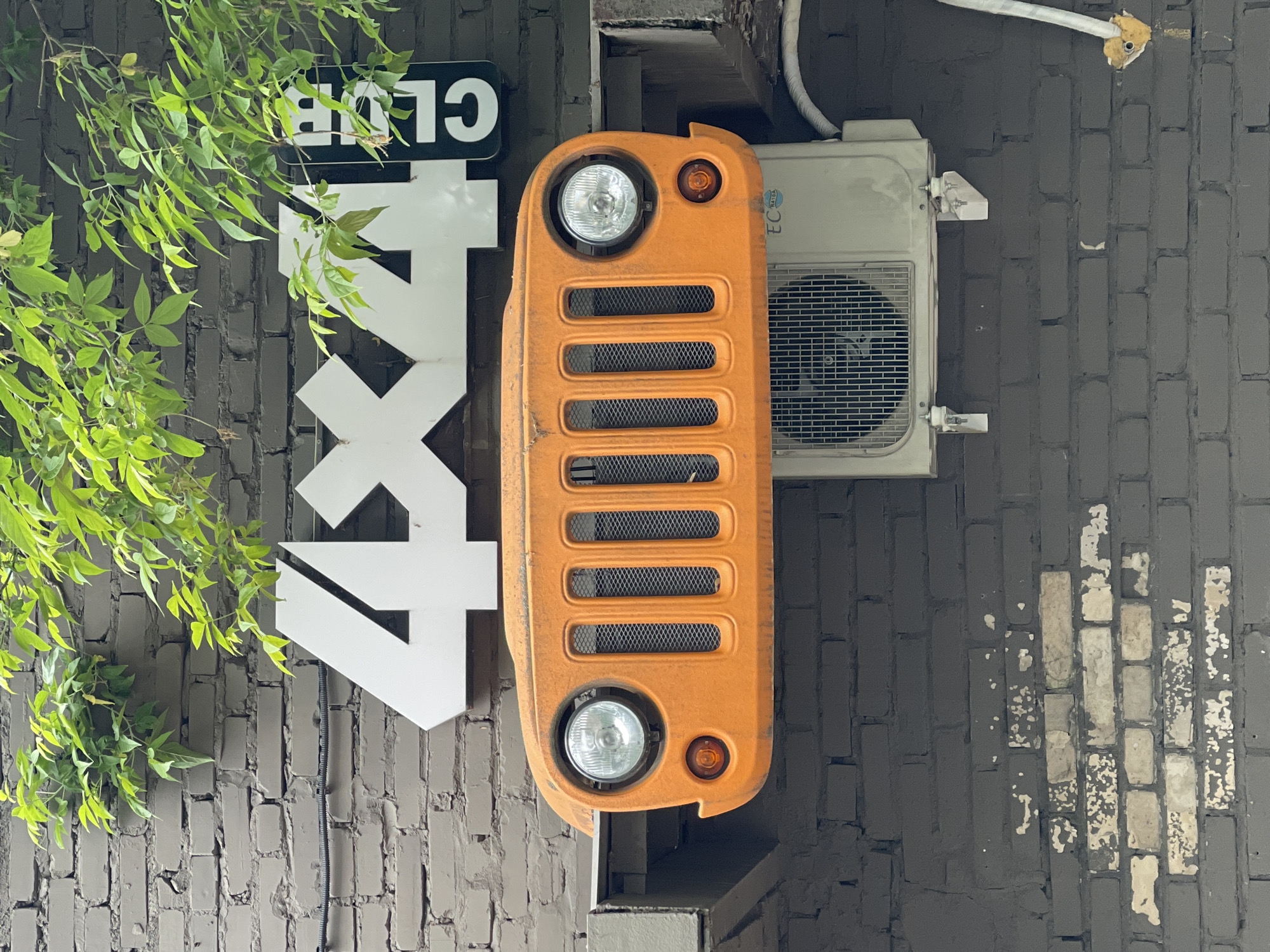 The image shows a section of a brick wall, possibly an exterior wall of a building. The wall is painted dark gray, with some bricks showing signs of wear or partial repainting. Mounted on the wall is a sign or decoration consisting of a stylized white "4x4" and "CLUB" lettering, partially obscured by green foliage. Next to the sign is an orange grille, likely a decorative piece resembling the front of a car, with headlights and indicator lights. Adjacent to the grille is a window air conditioning unit mounted on the wall. The location is identified as Khimki, Russia.