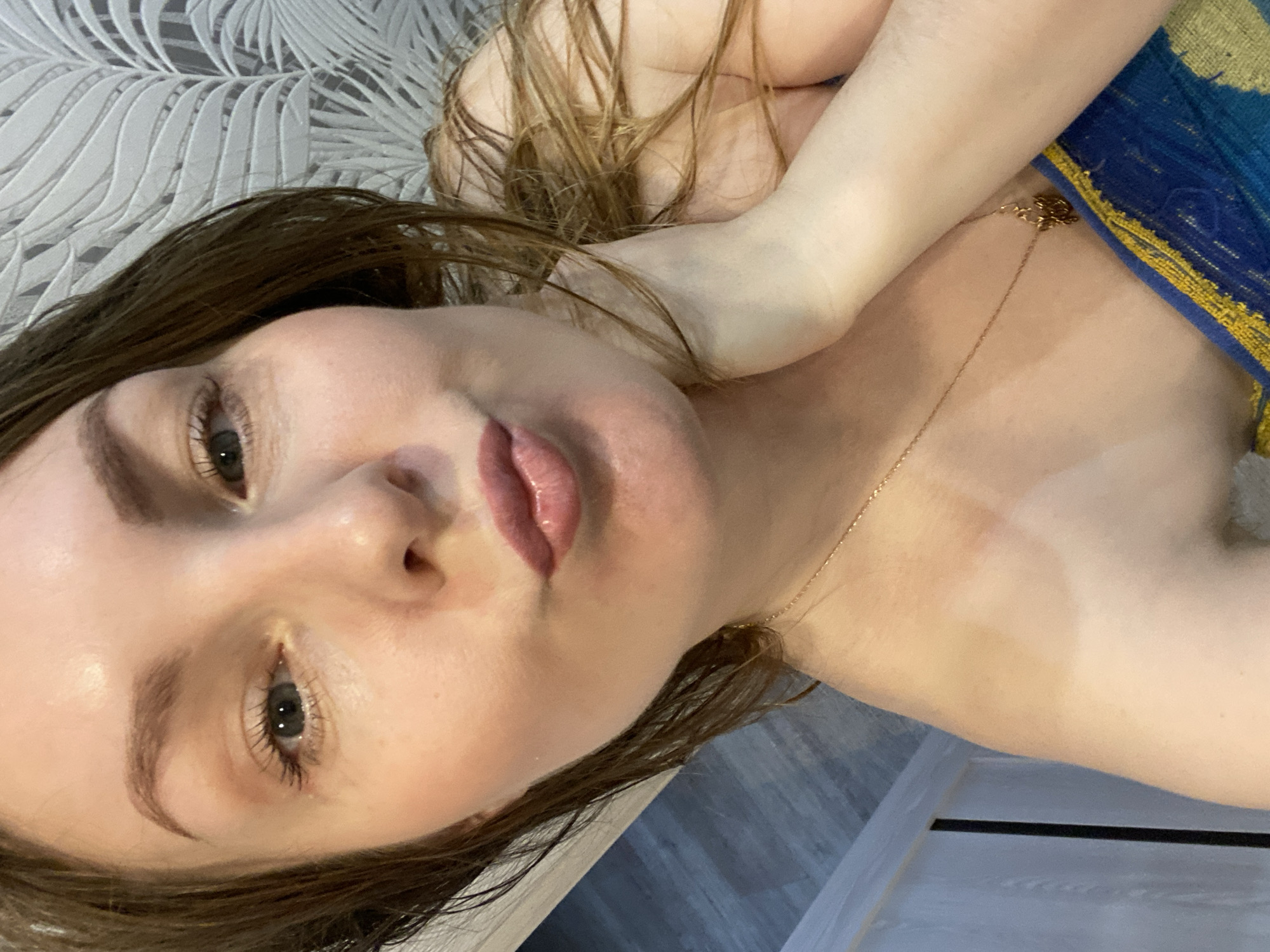 A young woman with light skin, brown hair, and a gold chain necklace is lying down. She is wearing a blue and yellow garment on her right shoulder. The background is a patterned wall and a gray floor. The city and specific location details are not discernible from the image.