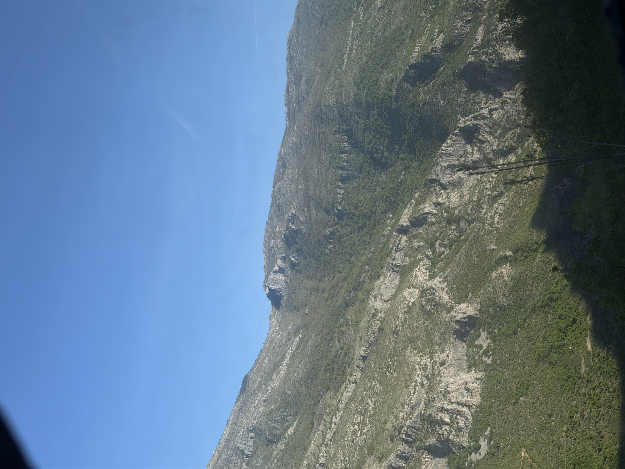 The image shows a mountain in Pelev Brijeg, Montenegro, with a clear blue sky in the background. The mountain is covered in vegetation and rocky terrain. There are shadows and sunlight playing on the mountain's surface, highlighting its contours and textures.
