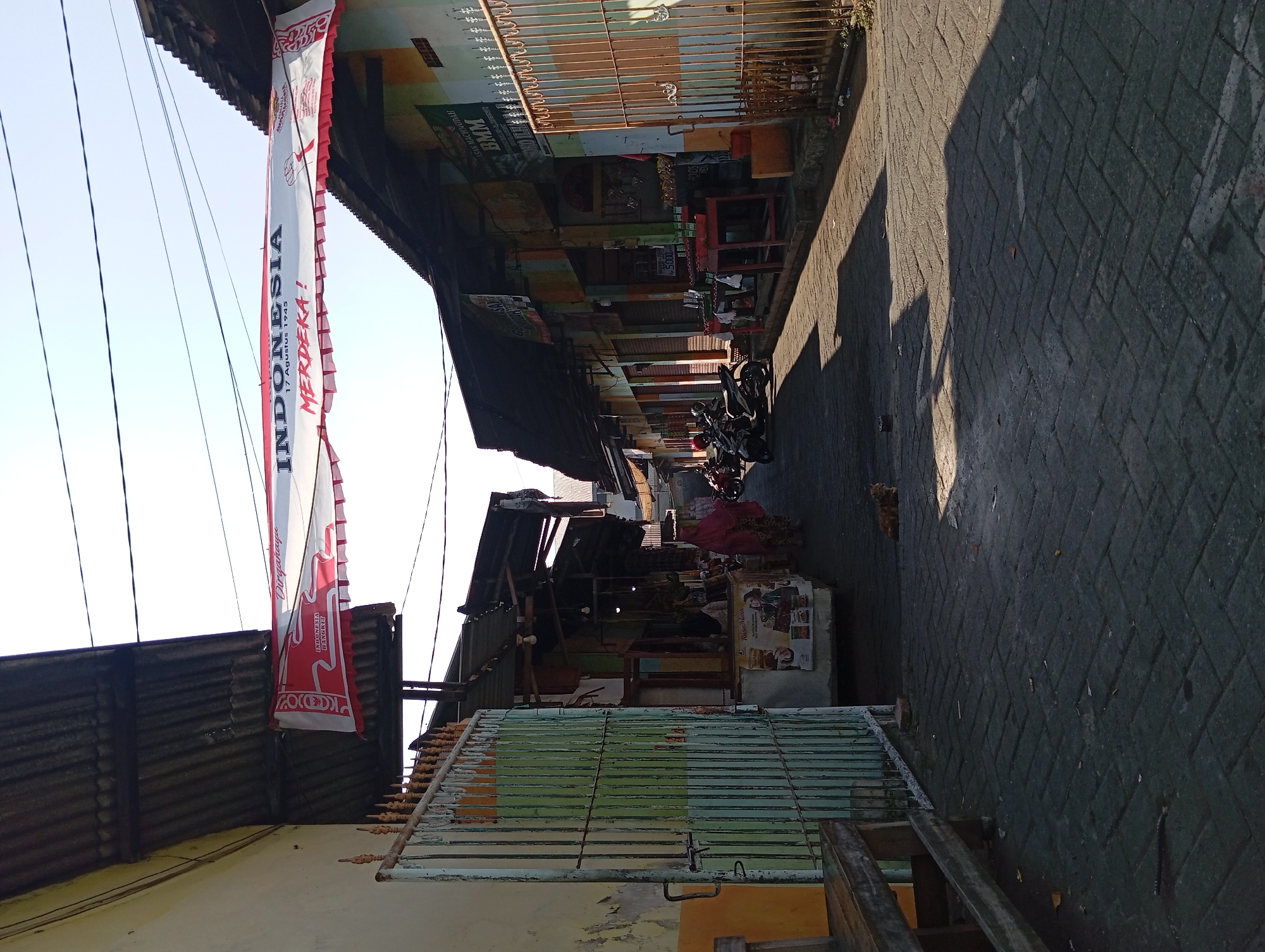 The image shows a street scene in the city of Kepulungan, Indonesia, likely in front of a market or commercial area. A banner is strung across the street, displaying the words "INDONESIA MERDEKA!" and the date "17 Agustus 1945," indicating a celebration of Indonesian Independence Day. The street is paved with dark grey interlocking bricks. On the left side of the street, there is a light green metal gate in front of a building with a peeling yellow and white facade, and a wooden bench is visible. On the right side, a row of shops with corrugated metal roofs and colorful facades line the street. Various items are displayed outside the shops, including what appear to be red shelves, bags, and some produce like bananas. Several motorcycles are parked along the right side of the street. A woman wearing a pink hijab and traditional clothing is walking away from the camera on the right side of the street, and a ginger cat is sitting on the brick pavement in the middle of the street, casting a shadow. The sun is shining, creating strong shadows from the buildings and objects in the scene. Overhead, power lines are visible against a clear blue sky.