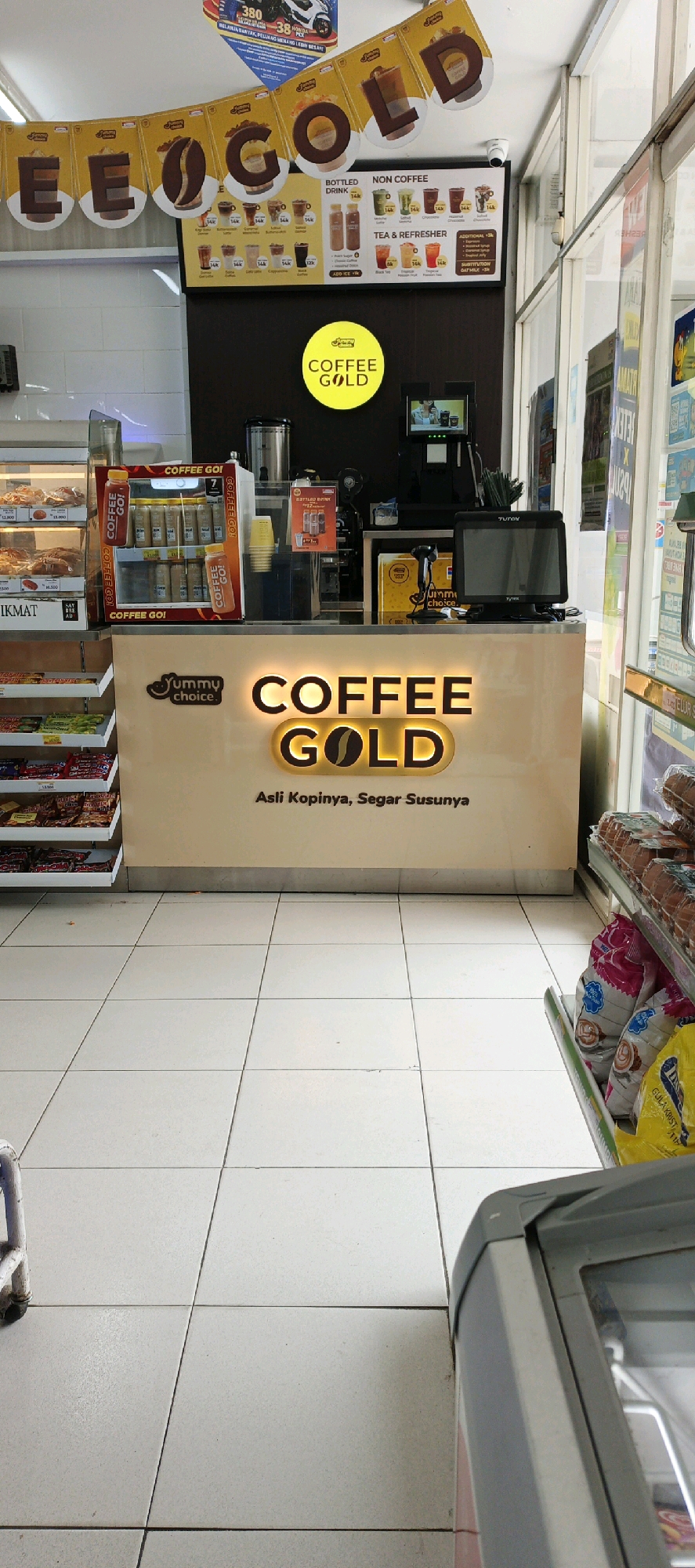 The image captures the interior of a convenience store, likely a minimarket, in Godean, Indonesia. The foreground is dominated by a tiled floor. In the mid-ground, a counter for a "COFFEE GOLD" stall is prominently displayed, featuring illuminated signage and a tagline in Indonesian that translates to "Its Coffee, Its Refreshing Milk." Behind the counter, various coffee-related products and equipment are visible, including bottled drinks in a refrigerated display, a coffee machine, and a point-of-sale system with a monitor. A menu board listing "BOTTLED DRINK," "NON COFFEE," and "TEA & REFRESHER" options is also visible, with prices indicated. The background reveals shelves stocked with packaged goods, including snacks and beverages. A banner overhead spells out "COFFEE GOLD." The overall impression is of a functional retail space offering coffee and convenience items.