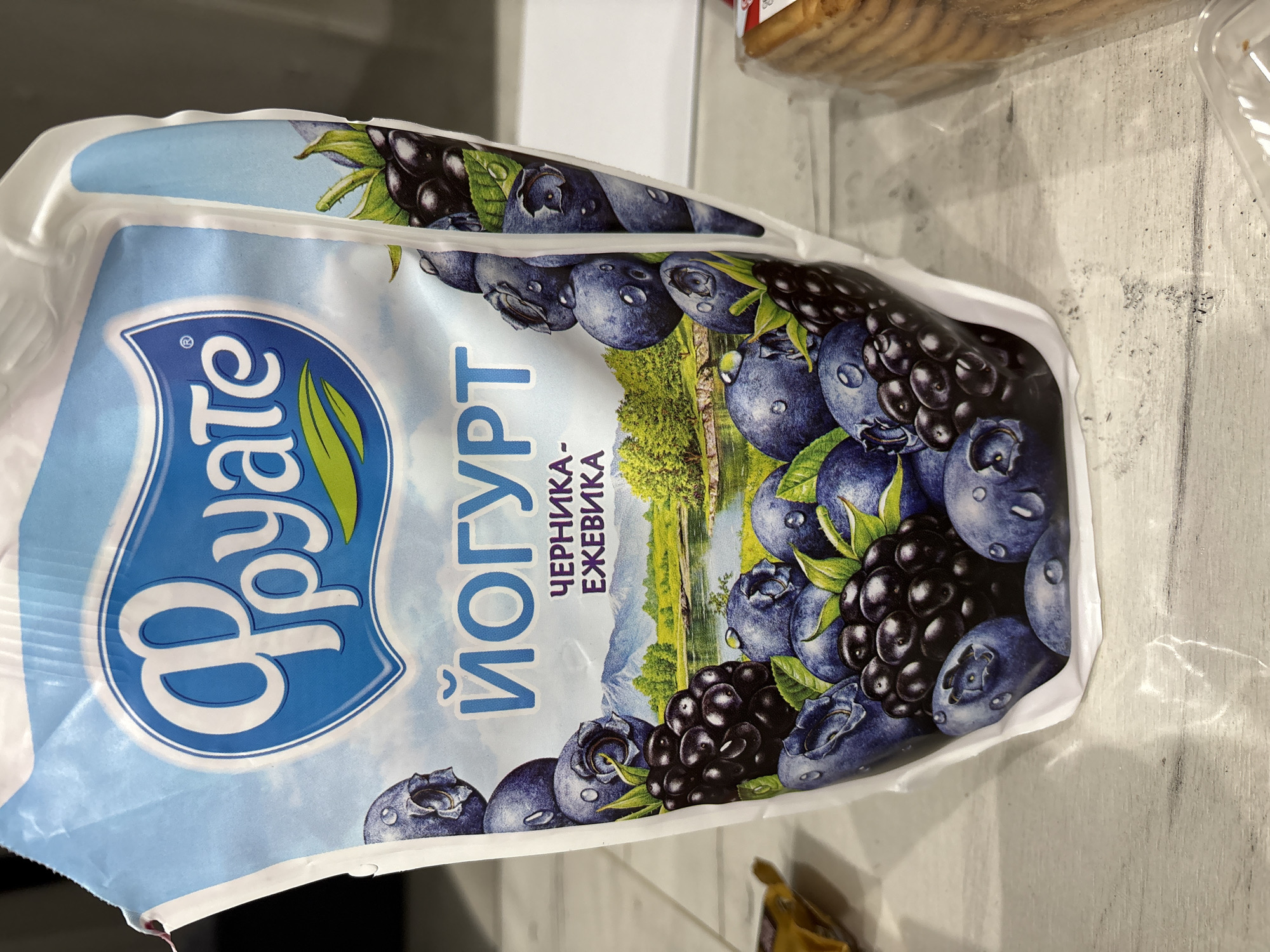 The image shows a package of "Фруате" (Fruate) yogurt with blueberry and blackberry flavor. The package is white with a blue background. The brand logo "Фруате" is in a blue shield-like shape with white lettering and green leaves. The word "Йогурт" (Yogurt) is written in blue Cyrillic next to the logo, and below that is the text "Черника-Ежевика" (Chernika-Ezhevika), which translates to Blueberry-Blackberry. The package also features an image of blueberries and blackberries, with a landscape in the background. The package is on a white surface. The location in the city of Unknown, Unknown is unidentifiable.
