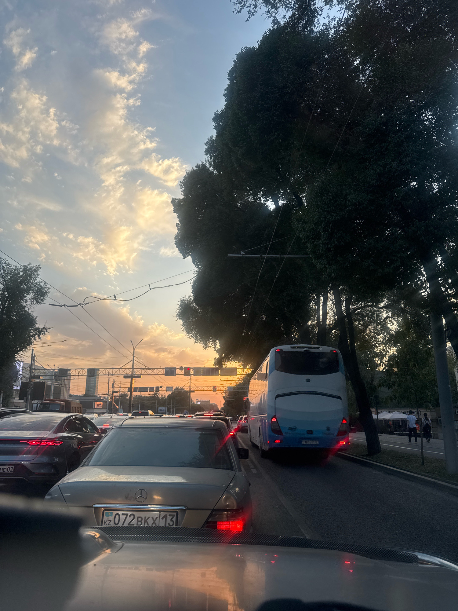 The image depicts a traffic jam in the city of Almaty, Kazakhstan, during sunset. A gold-colored Mercedes-Benz sedan with a license plate that reads "KZ 072BKX 13" is in the foreground, facing away from the camera. Behind it, a white and blue bus is visible, also in traffic. Several other cars are present, indicating heavy congestion. The sky is filled with dramatic clouds illuminated by the setting sun, casting a warm glow over the scene. Tall trees line the right side of the road, partially obscuring the sky. The overall environment suggests an urban setting with a street lined with traffic lights and power lines.