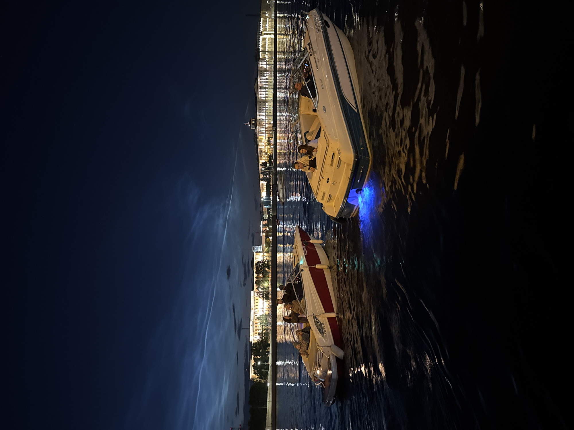 Two motorboats are navigating a dark river at night. The boat on the left is white with red trim and carries five people, two of whom are in the front and three in the back. The boat on the right is white with blue trim and carries three people; two are seated in the back and one is at the helm. The boat on the right has blue underwater lights. In the background, a large, illuminated building complex stretches along the riverbank, its lights reflecting on the water. The sky above is dark blue with wispy, luminous clouds. The scene is likely Saint Petersburg, Russia, possibly on the Neva River, given the distinctive architecture of the buildings and the presence of noctilucent clouds, which are often visible in northern latitudes during summer.