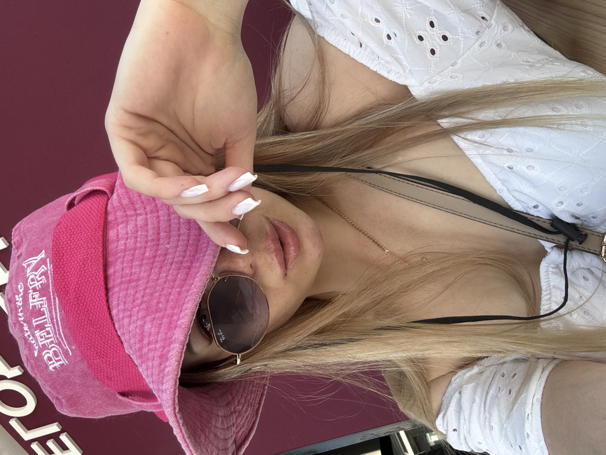 A young woman is seen in a close-up, waist-up shot, wearing a bright pink bucket hat and sunglasses that partially obscure her face. Her hand is raised, covering part of her eyes and forehead. She is also wearing a white, eyelet-detailed off-the-shoulder top and a delicate gold necklace. Her blonde hair is visible, falling over her shoulders. The background is a deep burgundy color. The image appears to be taken outdoors on a sunny day, possibly in an urban setting given the text visible on the wall in the upper left corner, which reads in large, white letters, "ЕГОРО".