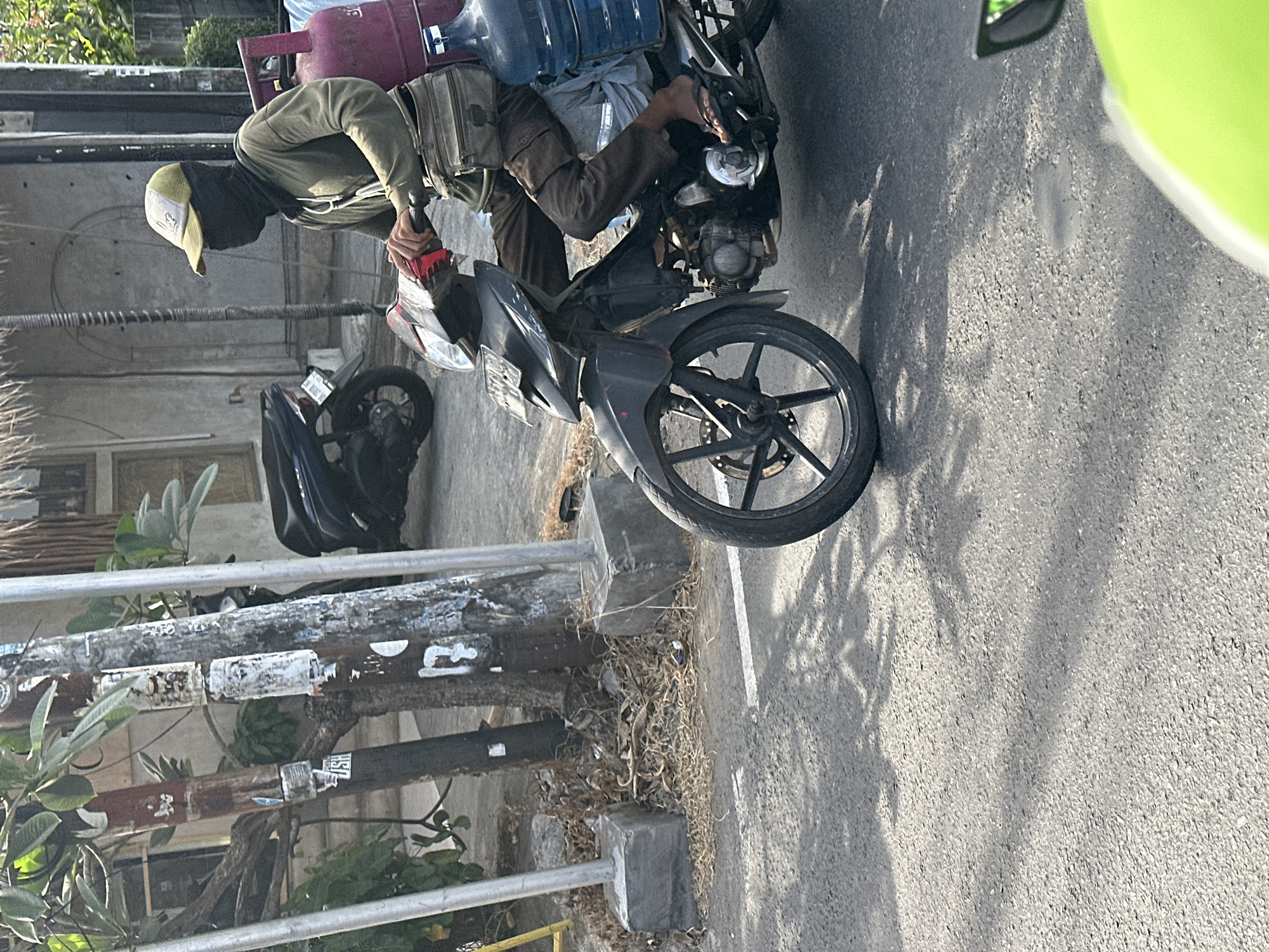 A person wearing a green long-sleeved shirt, brown pants, a yellow and green baseball cap, and a black face covering is riding a dark-colored motorcycle on a paved road. The person is carrying a large pink propane tank and a large blue water jug on the back of the motorcycle. The scene is outdoors in North Kuta, Indonesia, with trees and buildings visible in the background. The road appears to be a street in a commercial or residential area, with shadows from trees cast on the asphalt.