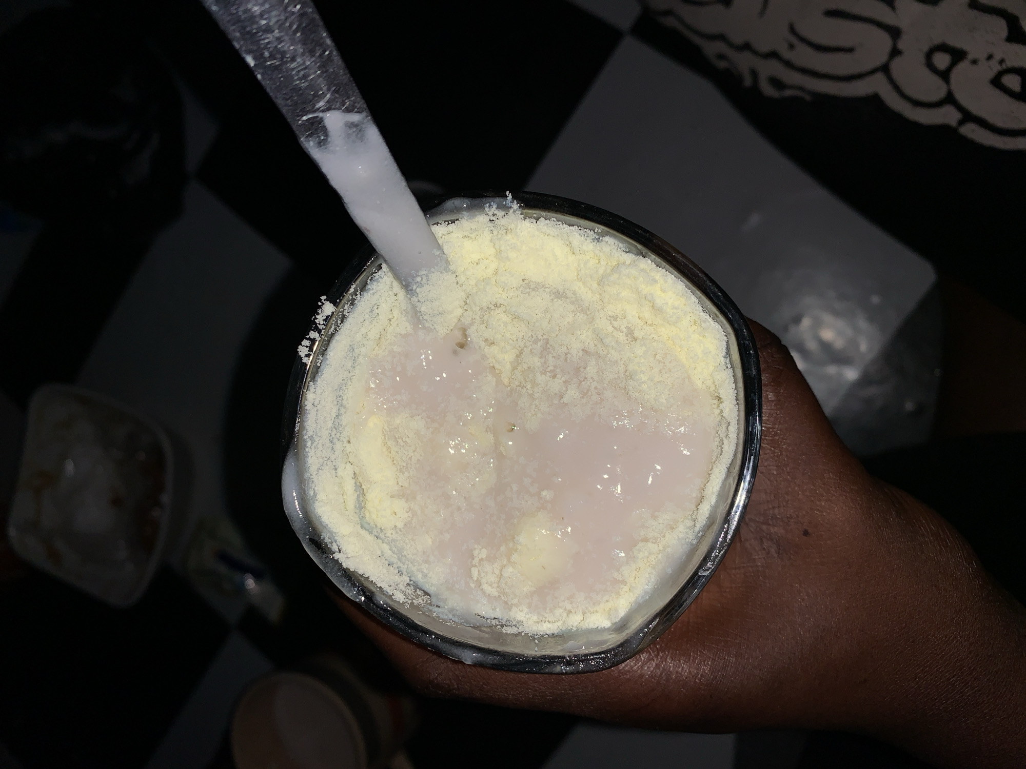A top-down shot reveals a glass of liquid, appearing to be a white drink with a layer of yellow powder on top, being stirred with a clear spoon or straw. The glass is held in a dark-skinned hand. The background features a black and white checkered surface and other indistinct objects. The location is Rafin Par, Nigeria, based on the prompt.