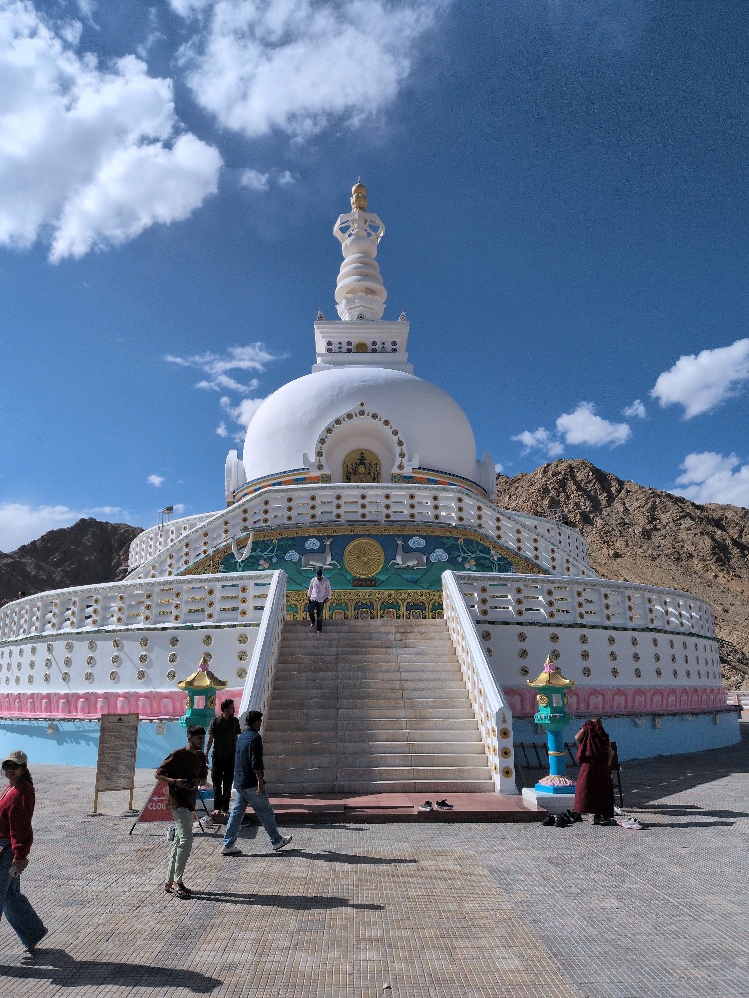 The image depicts the Shanti Stupa, a Buddhist white-domed stupa located on a hilltop in Leh, Ladakh, India. The stupa is a prominent landmark, featuring intricate decorative bands and a golden spire. In front of the stupa, a wide staircase leads up to its entrance. Several individuals are visible in the scene. On the stairs, a person in a pink shirt is walking up. On the plaza in front of the stupa, a group of people are seen. To the left, a woman in a red sweater and jeans is walking away from the camera. Near the center, three men are standing and walking. To the far right, a person in a maroon robe, possibly a monk, is standing near a decorative pillar. The ground is paved with patterned tiles, and shadows are cast by the people and structures. In the background, arid, rocky hills are visible under a bright blue sky with scattered white clouds.