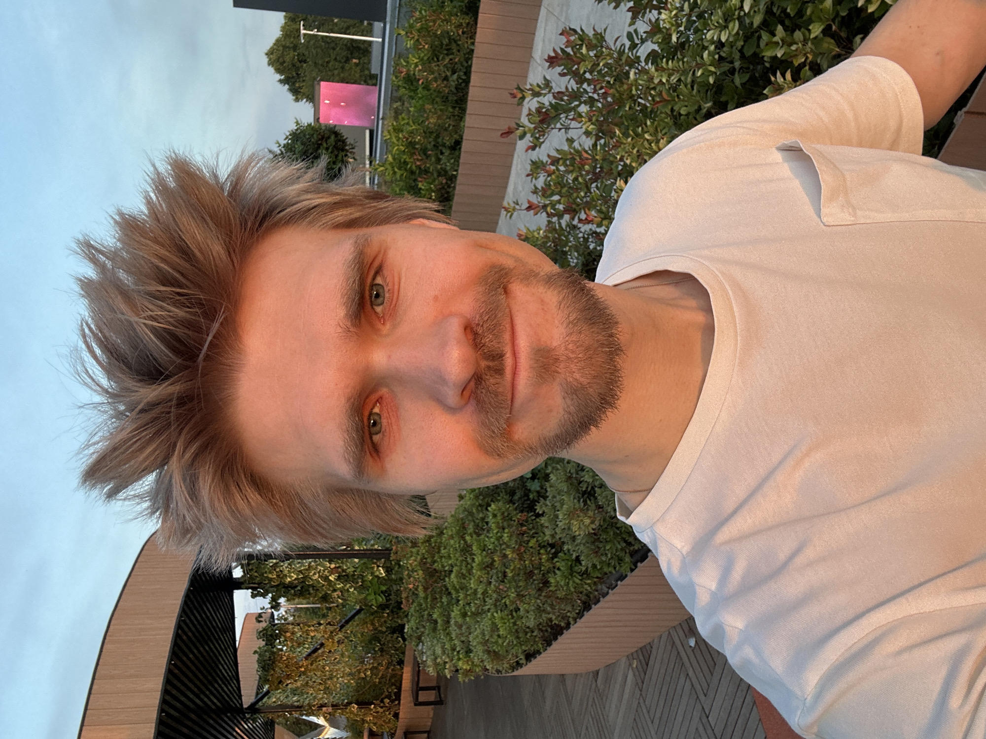 A photograph depicts a fair-skinned male with light blond hair and a beard stubble. He is wearing a white T-shirt with a pocket and is positioned slightly angled to the left. The background features outdoor foliage including bushes and small trees, likely on a rooftop. In the far background, a tall structure is visible, as well as a pink sign. The location is indicated as being in Tashkent, Uzbekistan.