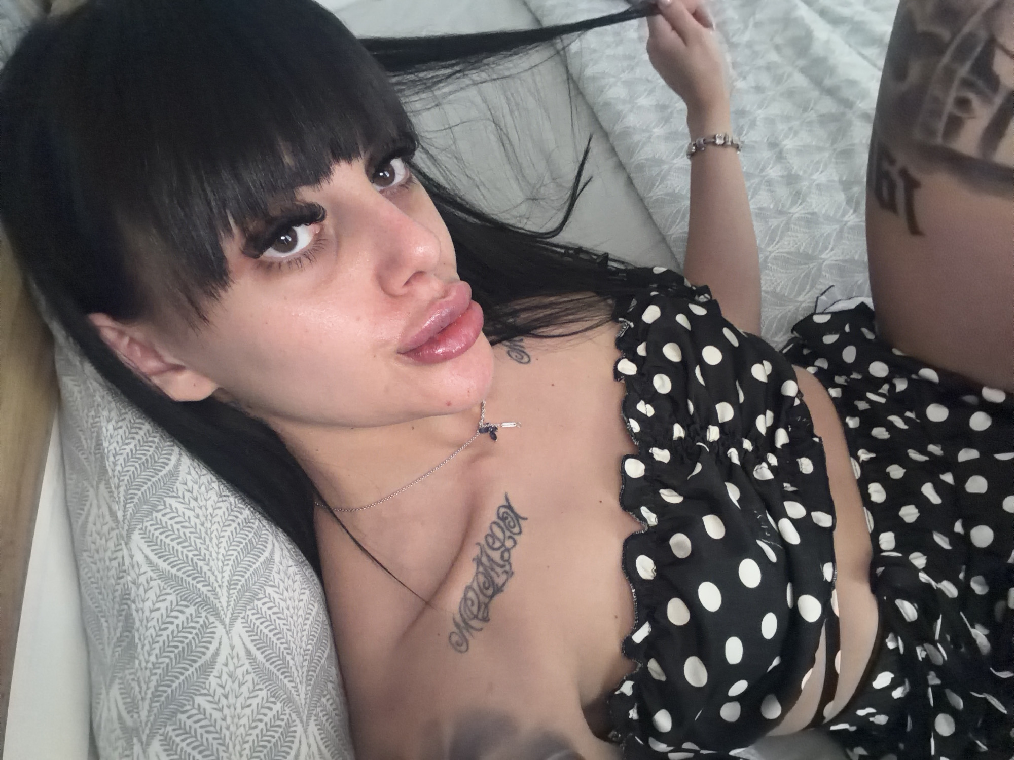 A young woman with long, dark hair and bangs lies on a bed with a patterned duvet. She is wearing a black and white polka dot crop top and skirt. She has several tattoos, including one on her chest and another on her thigh. Her makeup includes false eyelashes and lip gloss. She is looking at the camera and holding a strand of her hair. The scene takes place indoors.