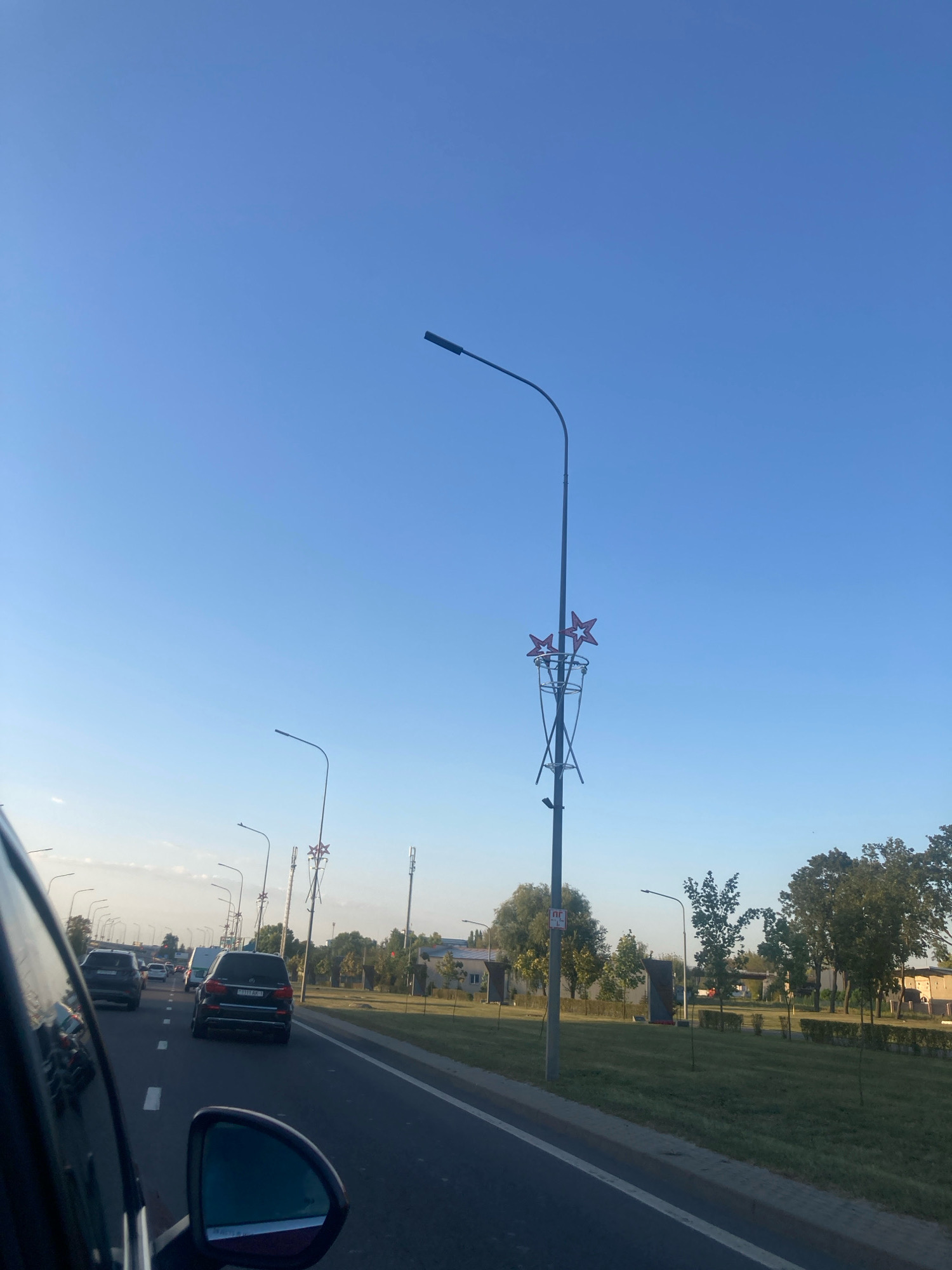 The image captures a view from a moving vehicle on a clear, sunny day, likely on a road leading into or out of the city of Brest, Belarus. The road is asphalt with visible white lane markings. Several cars are in motion, including a black SUV in the foreground. Along the right side of the road, there is a grassy median with trees and lampposts. One lamppost features a decorative element with three red stars. In the background, buildings and more trees are visible under a vast, pale blue sky. The overall scene depicts a typical urban or suburban roadside environment.