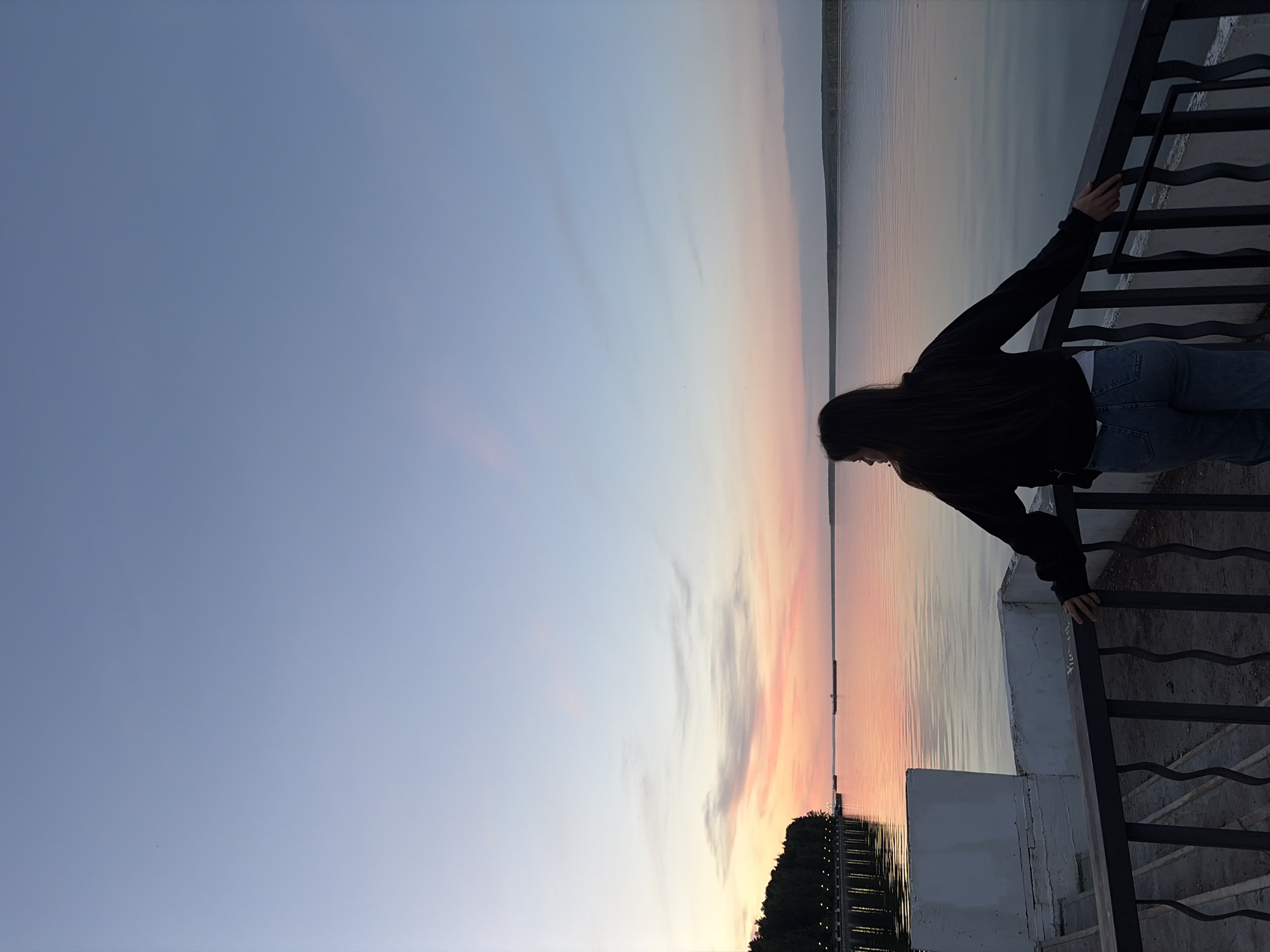 The image, taken in Chaikovsky, Russia, captures a serene moment at either sunrise or sunset over a vast body of water, likely the Kama River or a reservoir. The scene is dominated by a clear sky displaying a beautiful gradient of deep blue at the top, transitioning to lighter blues, soft pinks, and warm oranges closer to the horizon, where the sun's low angle creates a picturesque glow. Wispy clouds are visible in the lighter parts of the sky.
In the foreground, a lone individual, a female with long, dark hair, is seen from behind and slightly to the side. She is wearing a dark long-sleeved jacket or hoodie over a lighter top and blue jeans. Her arms are resting on a dark metal railing, which features decorative, wavy patterns, as she gazes thoughtfully across the tranquil water. The railing is supported by a white, concrete or stone balustrade.
The calm water surface perfectly reflects the colors of the sky, creating a mirroring effect that enhances the overall peaceful atmosphere. In the distance, on the opposite bank, a thin strip of land or trees is visible, silhouetted against the bright horizon. Along the lower left edge of the image, the shoreline appears to have a line of lights, suggesting an urban or semi-urban promenade. The overall lighting is dimming, typical of dawn or dusk. On the white balustrade below the railing, there are some faint, indistinct markings or text, appearing like graffiti. The image conveys a sense of calm contemplation as the individual takes in the expansive view during the golden hour.