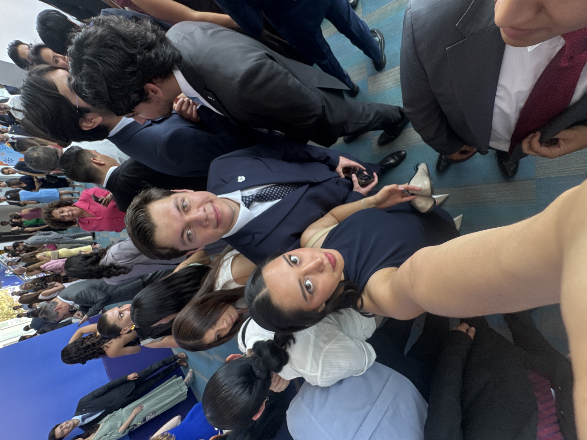 This image appears to be a selfie taken at an event, likely a graduation or similar formal gathering, in Puebla, Mexico. A group of young adults are crowded together, with the camera held at an angle to capture as many people as possible. Many of the individuals are wearing formal attire, such as suits and dresses. There are indications of a structured event, with many people sitting or standing closely together. The location appears to be indoors on a carpeted floor. The general atmosphere seems celebratory and communal.