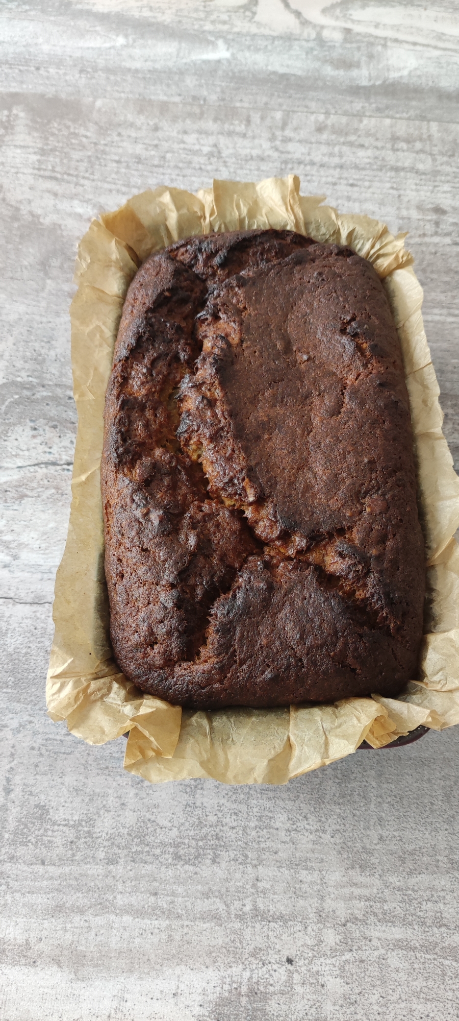 The image shows a close-up, overhead view of a loaf of dark brown, baked goods, likely bread or cake, presented in a baking dish lined with crinkled parchment paper. The loaf has a rustic, uneven texture with a crack on the top surface, indicating it has just been baked. The parchment paper extends beyond the edges of the baking dish, creating a soft, textured border. The background is a light grey, textured surface, possibly a wooden table or countertop, with visible grain patterns and a slightly distressed appearance. The lighting is natural and diffused, casting subtle shadows. The scene is stationary and does not depict any people or actions. The location is not specified in the image itself, and therefore, cannot be determined to be in Rosinka, Russia.