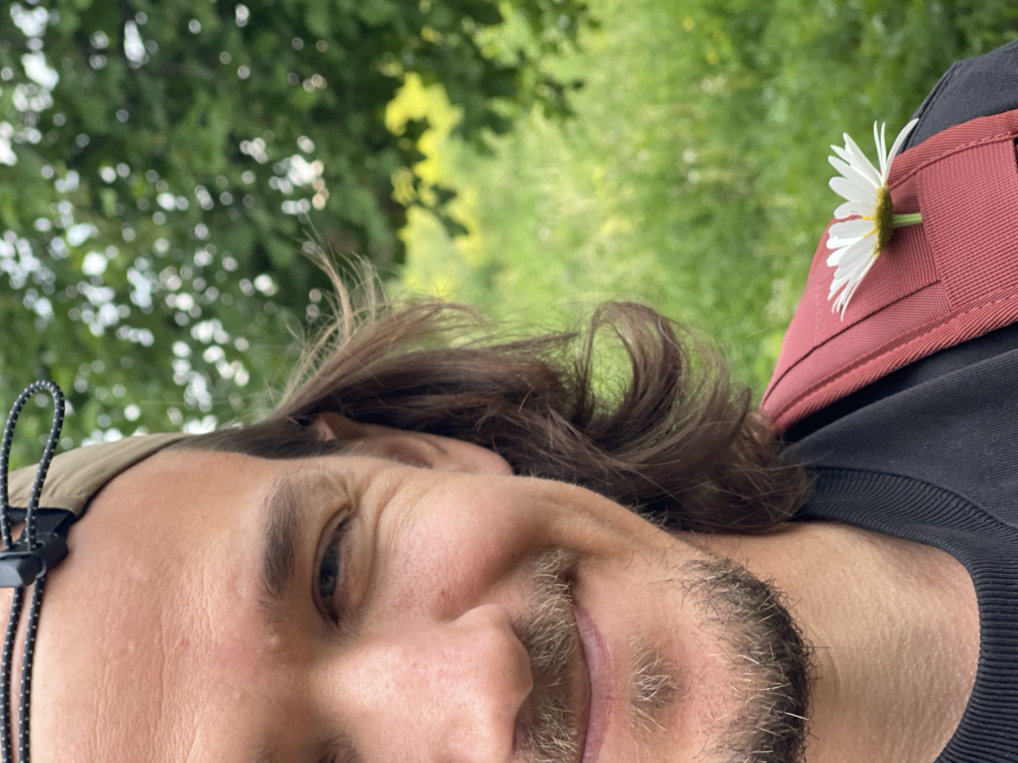 A close-up selfie shows the right side of a person's face and upper torso. The person has brown hair, a mustache, and a goatee. They are wearing a black t-shirt and a tan baseball cap with a black cord. A red backpack strap is visible on their right shoulder, with a white daisy tucked into it. The background is blurred green foliage, suggesting an outdoor, natural setting. The image is taken from a low angle, looking slightly up at the subject.