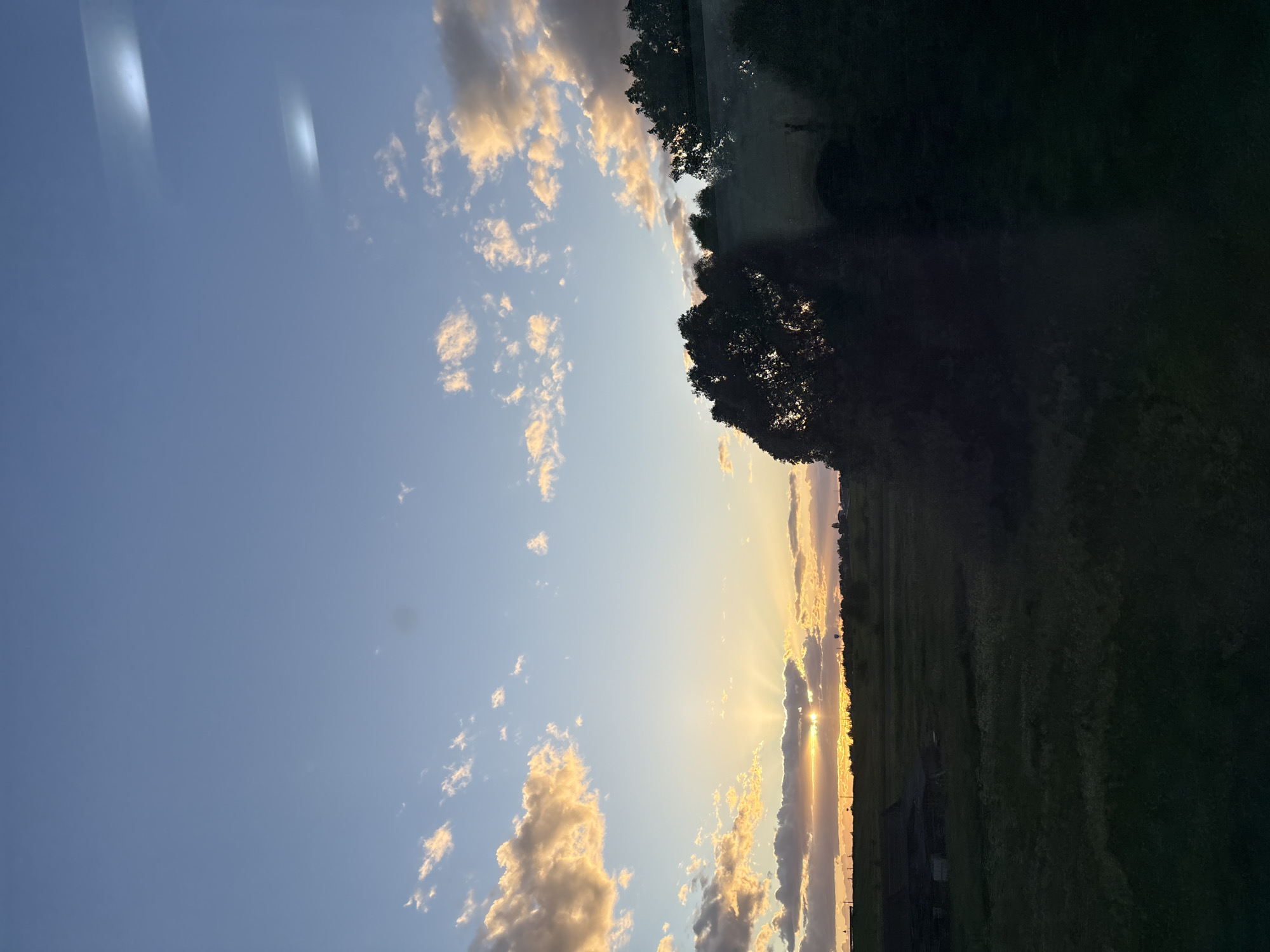 The image captures a sunset scene in Мажэйкаўскі сельскі Савет, Belarus. The sky is a gradient of blue, transitioning to orange and yellow as it nears the horizon, with scattered cumulus clouds illuminated by the setting sun. Sunlight streams through a gap in the clouds, creating rays that extend downwards. Along the horizon, trees and vegetation silhouette against the bright light. The foreground consists of a dark, undefined area, potentially a field or wooded area. There is no presence of people.
