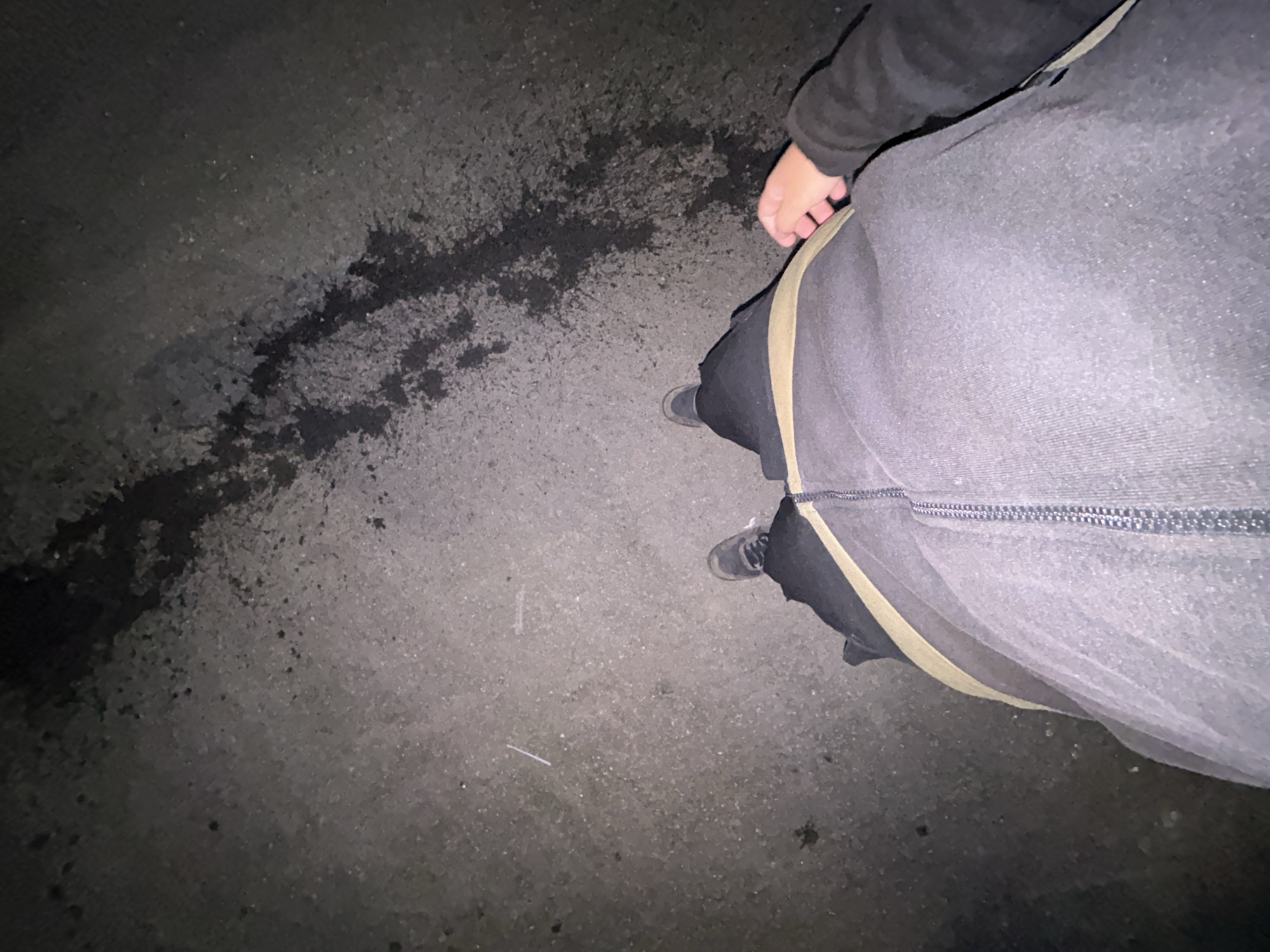 The image shows a first-person perspective of a person standing on an asphalt surface, likely a road or sidewalk, at night. The ground is dark with some lighter speckled areas and a dark, wet-looking stain in the upper portion of the frame. The person is wearing dark pants, a light-colored belt or waistband, and dark shoes. A hand with visible fingers is resting on the person's thigh. The lighting suggests an urban environment, and the detail about the location of Gorna Malina, Bulgaria, is not visually inferable from the image itself.