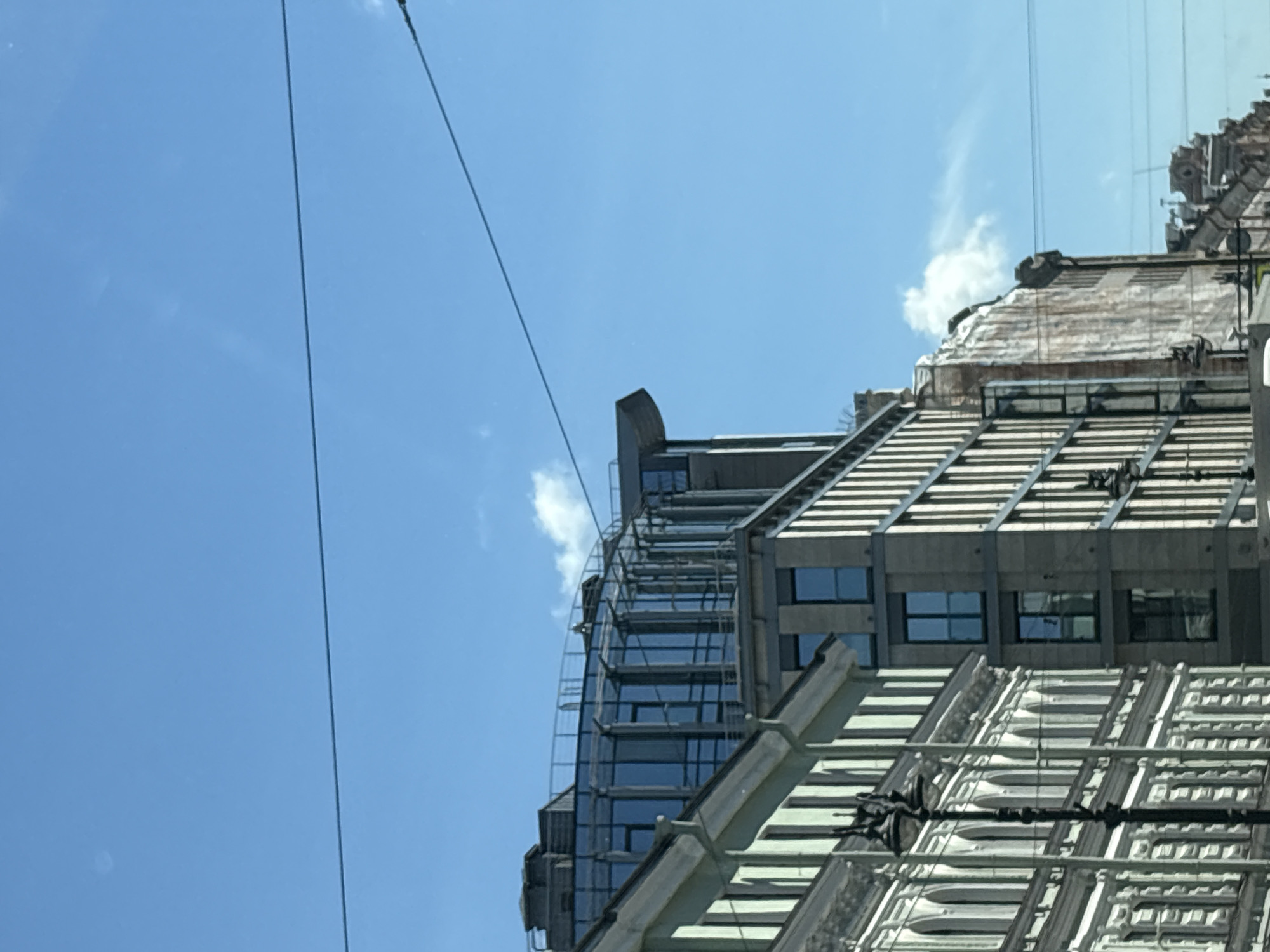 The image captures a clear blue sky above a street in Saint Petersburg, Russia, with several buildings lining the thoroughfare. On the left, a classical building with ornate white detailing and arched windows is visible. Adjacent to it is a modern, multi-story building with a facade composed of numerous windows and vertical accents. Further down the street, another building displays a weathered, textured surface, and beyond that, more urban architecture with visible rooftop elements. Power lines are strung across the sky, adding a common urban element to the scene. The lighting suggests it is daytime, with sunlight illuminating the facades of the buildings.