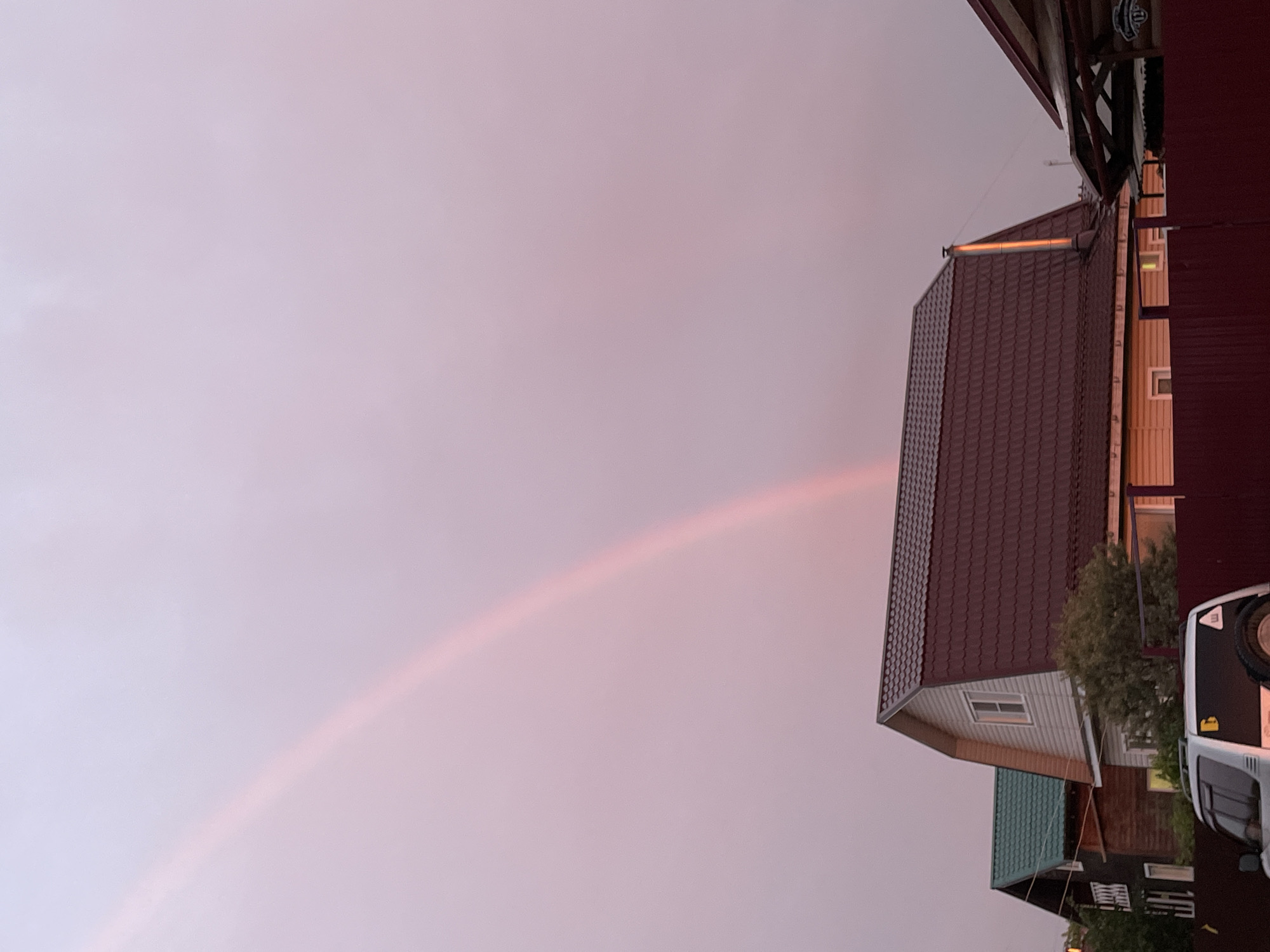 The image depicts a cloudy sky with a subtle pink hue, possibly indicating sunset or sunrise. A faint rainbow arc is visible stretching across the upper portion of the frame. In the foreground, several houses with distinct roofing styles are present. The house in the center has a dark red, tiled roof and light-colored siding. To its left, another house features a green, tiled roof. A dark red fence runs along the bottom of the image, and a white SUV is parked in front of it. The overall scene suggests a suburban or rural setting within the city of Severodvinsk, Russia.