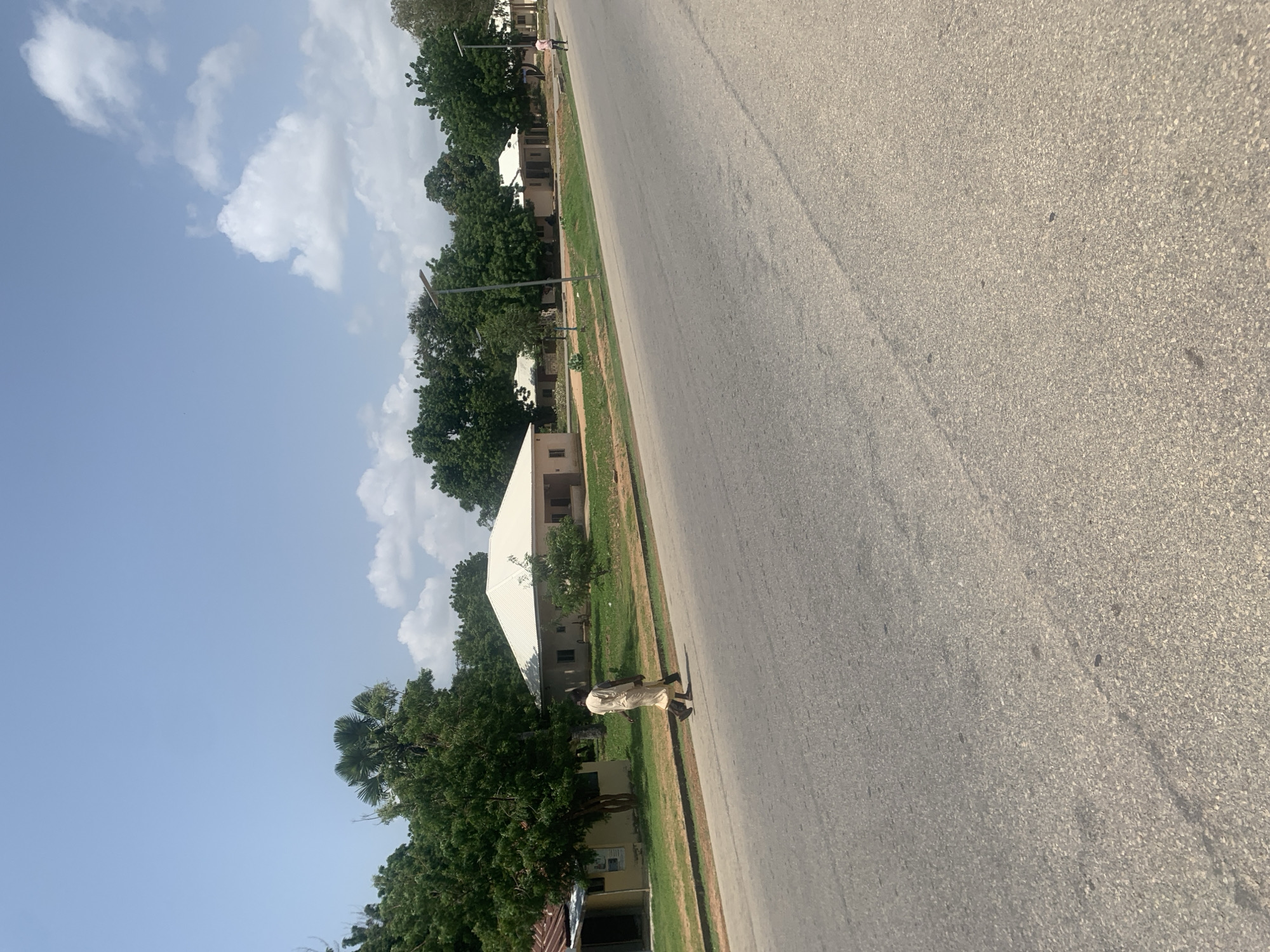 The image shows a street scene in Jalingo, Nigeria. A person in light-colored clothing is walking on the right side of the paved road. The person appears to be male, wearing a tunic and trousers. The road stretches diagonally across the frame, with a grassy median to the left. Along the median, there are trees and some buildings with white roofs. In the background, more trees and buildings are visible. The sky is blue with scattered white clouds. There are also lampposts along the side of the road. The lighting suggests it is daytime, likely in the morning or afternoon. The overall setting appears to be a residential or institutional area. There is no visible text in the image.