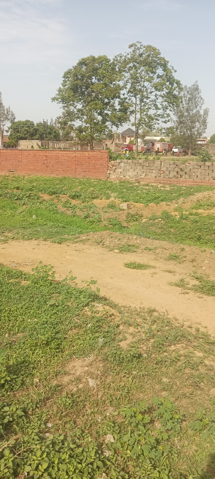The image shows a vacant lot in Jos, Nigeria. In the foreground is a dirt path with sparse green vegetation. A low wall made of red bricks and gray concrete blocks is in the midground. Behind the wall are several buildings and a couple of fire trucks with red paint. There are people standing behind the fire trucks. Two large green trees stand behind the wall and buildings. The sky is overcast.