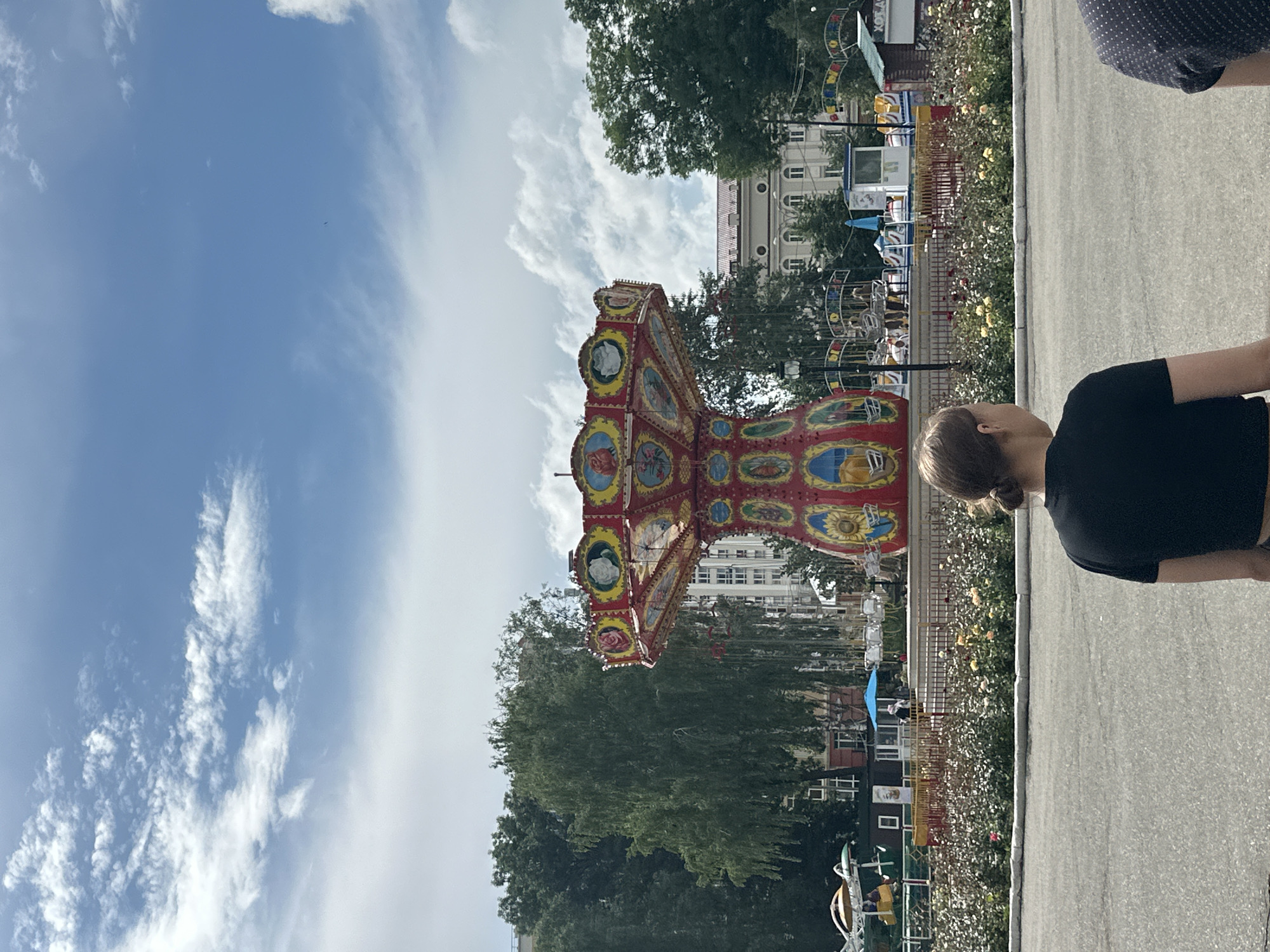 A overhead view of an amusement park in Saransk, Russia features a classic swing ride with red and yellow accents, decorated with circular paintings of flowers and faces. The ride stands prominently in the center of the frame, surrounded by lush green trees and a well-maintained flower bed blooming with yellow and pink roses. In the foreground, a young woman with her hair tied back in a bun, wearing a black t-shirt, faces away from the camera, looking towards the amusement park. The sky above is a bright blue, dotted with fluffy white clouds, suggesting a pleasant day. In the background, a few buildings are visible, some with intricate architectural details. The scene depicts a lively and colorful public space, likely a park or a fairground, on a sunny day.