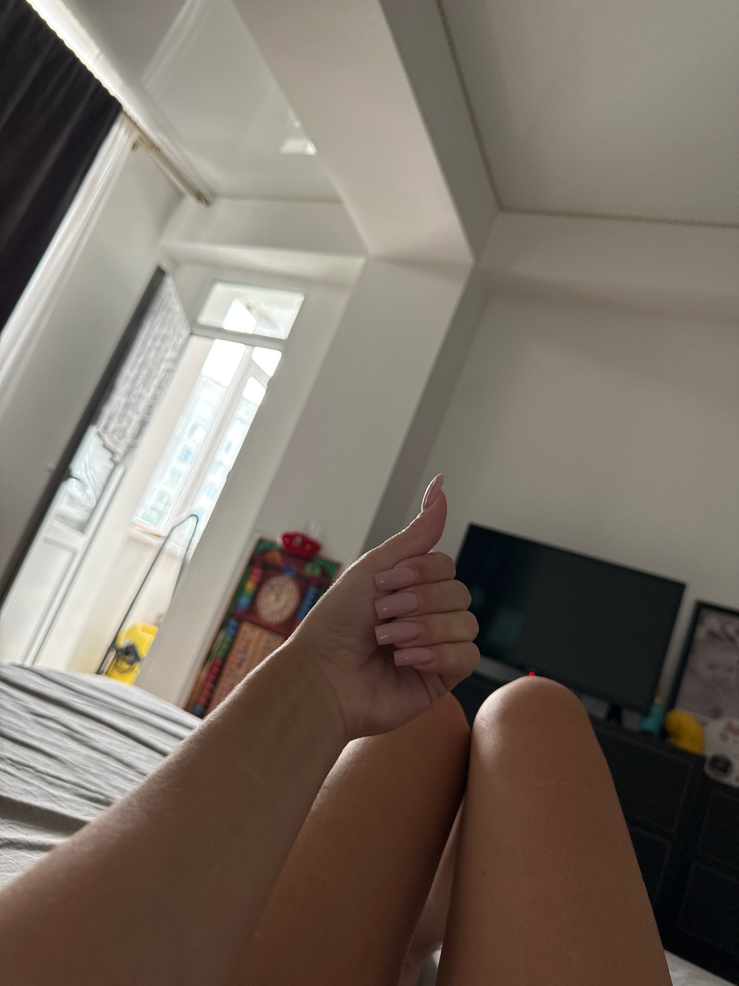The image is an indoor shot, likely a bedroom, with natural light streaming from a window. The camera is positioned to capture a low-angle view of a person's arm and legs, with a thumb raised in a "thumbs up" gesture. The arm is lightly tanned and shows some texture. The legs are also tanned. In the background, a dark television screen is visible, along with a black dresser. To the left, a window with blinds is visible, offering a view of a building with multiple floors and windows. A black curtain is partially obscuring the window. A colorful tapestry or artwork hangs on the wall near the window. The scene suggests a casual, relaxed setting. There is no indication of the city being Chișinău, Moldova.