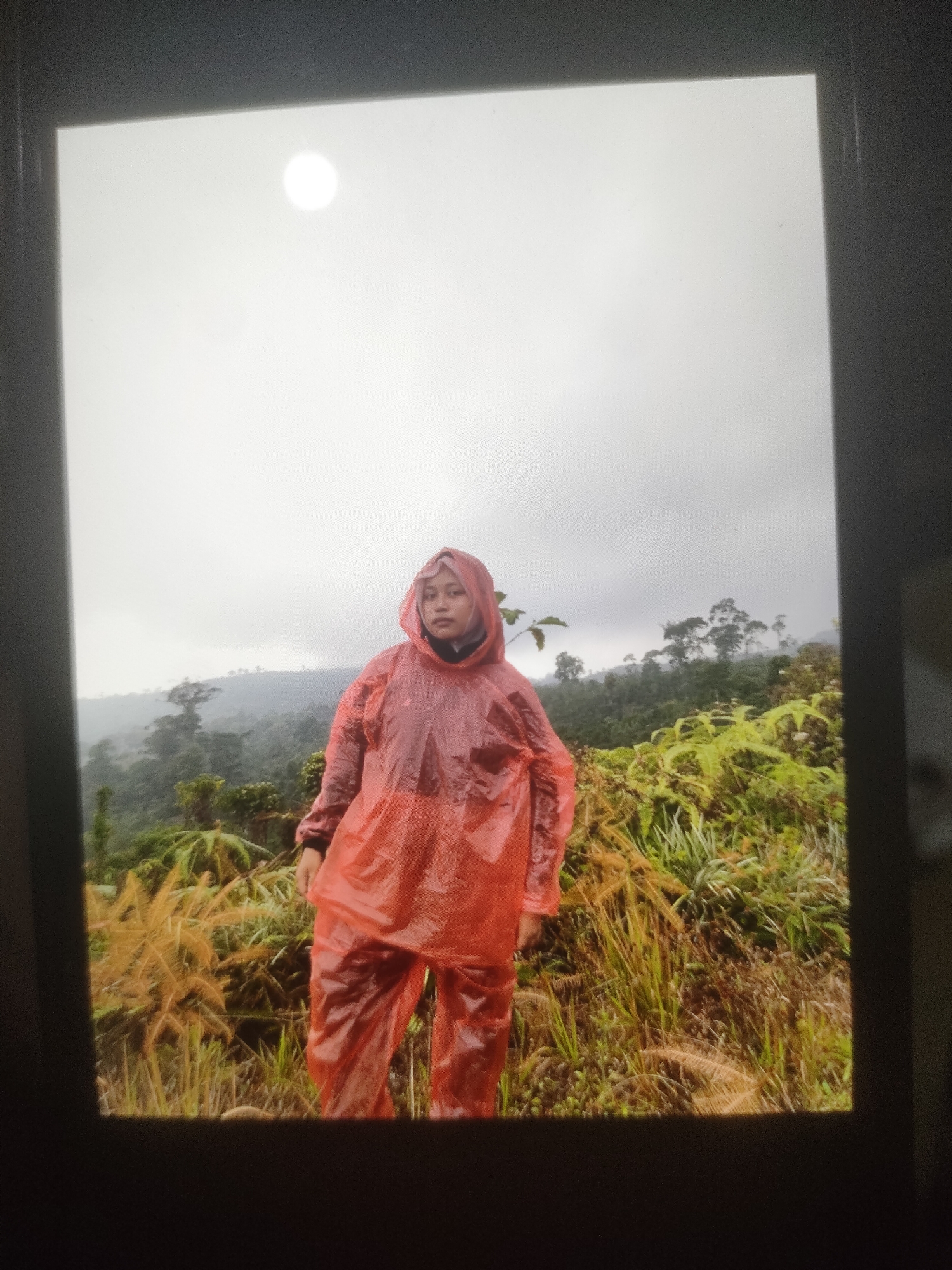 A young woman stands in a bright orange raincoat and pants amidst a lush, green landscape with rolling hills in the background. The sky is overcast, with a bright, diffused light source visible at the top left, possibly the sun obscured by clouds. The woman is wearing a hijab and appears to be looking directly at the camera. The foreground consists of tall, dry grasses and green foliage. The location is identified as Padang Dalom, Indonesia. The image captures a moment during what appears to be an outdoor excursion, possibly hiking or exploring, in a natural, somewhat misty environment.