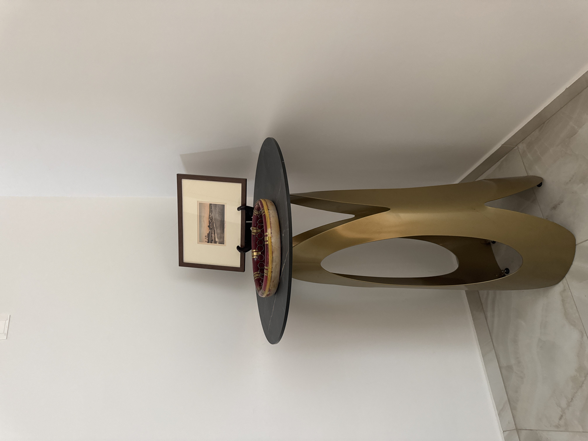An indoor shot captures a corner of a room in Germasogeia Municipality, Cyprus. The scene features a round, dark-topped table with a distinctive golden base. On the table, a framed picture rests on a black easel, and a decorative bowl containing red and yellow items is visible. The walls are painted white, and the floor is tiled with a marble-like pattern. The lighting suggests an interior environment.